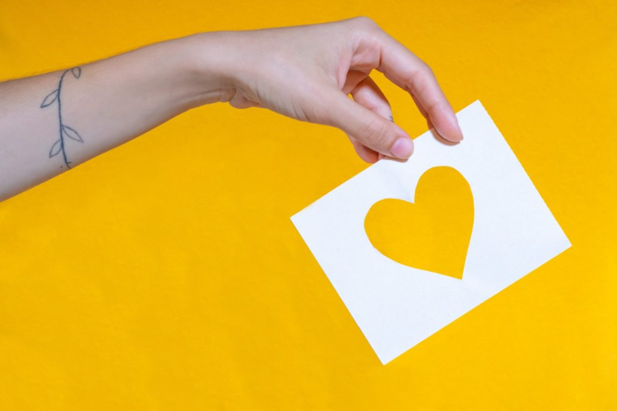 uma mão segura um papel cortado que abriga a forma de um coração. O fundo da foto é amarelo.