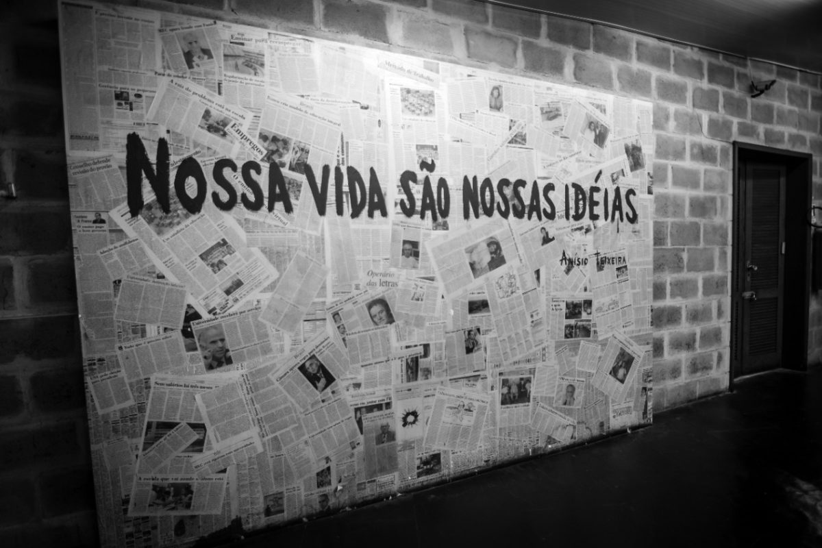 Em uma parede tijolos, há um mural com várias folhas de jornal. Sobre eles, escrito a tinta lê-se: "Nossa vida são nossas idéias". A foto está em preto e branco.