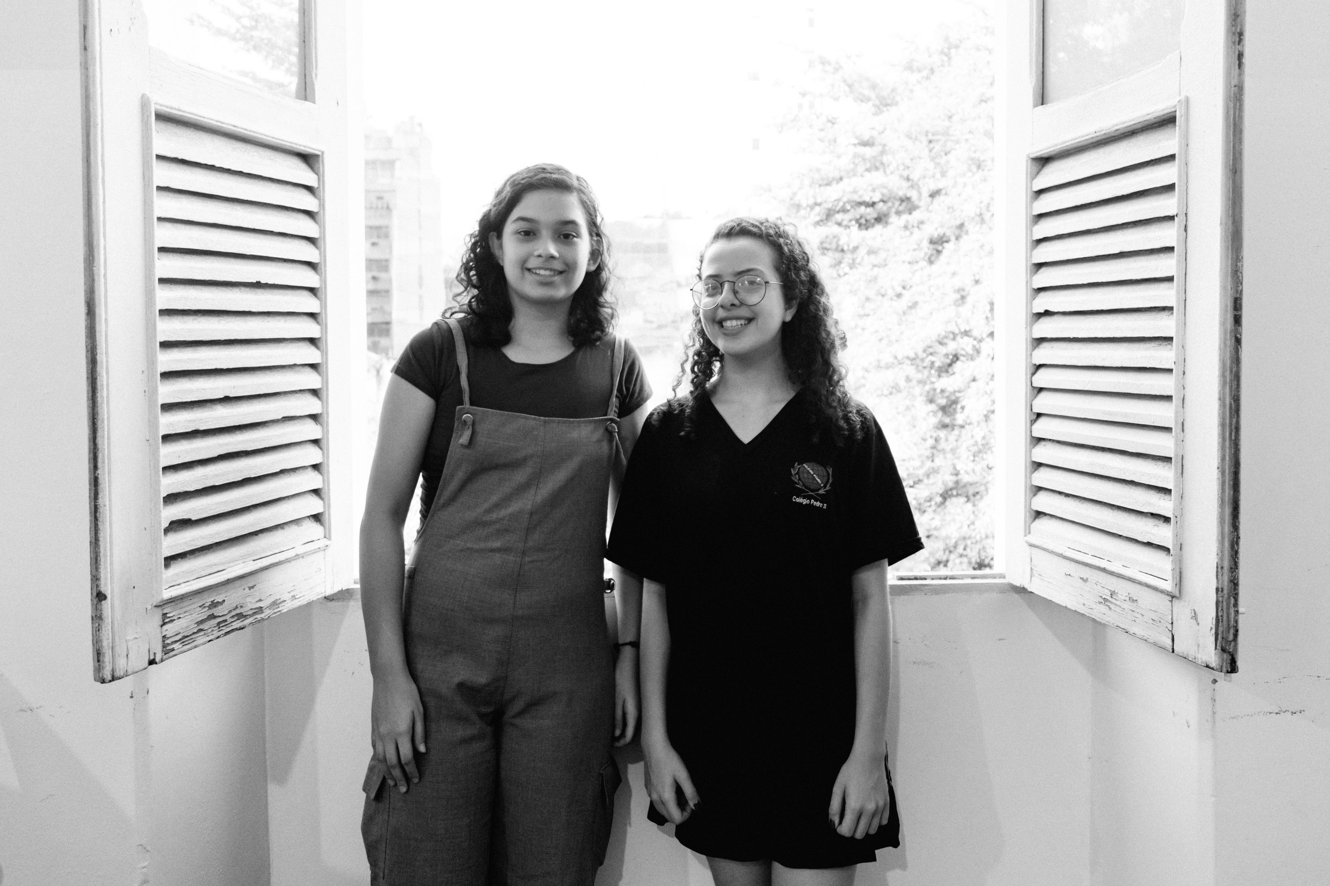 Gabriela e Catelina estão em frente à janela e sorriem para a foto, que está em preto e branco