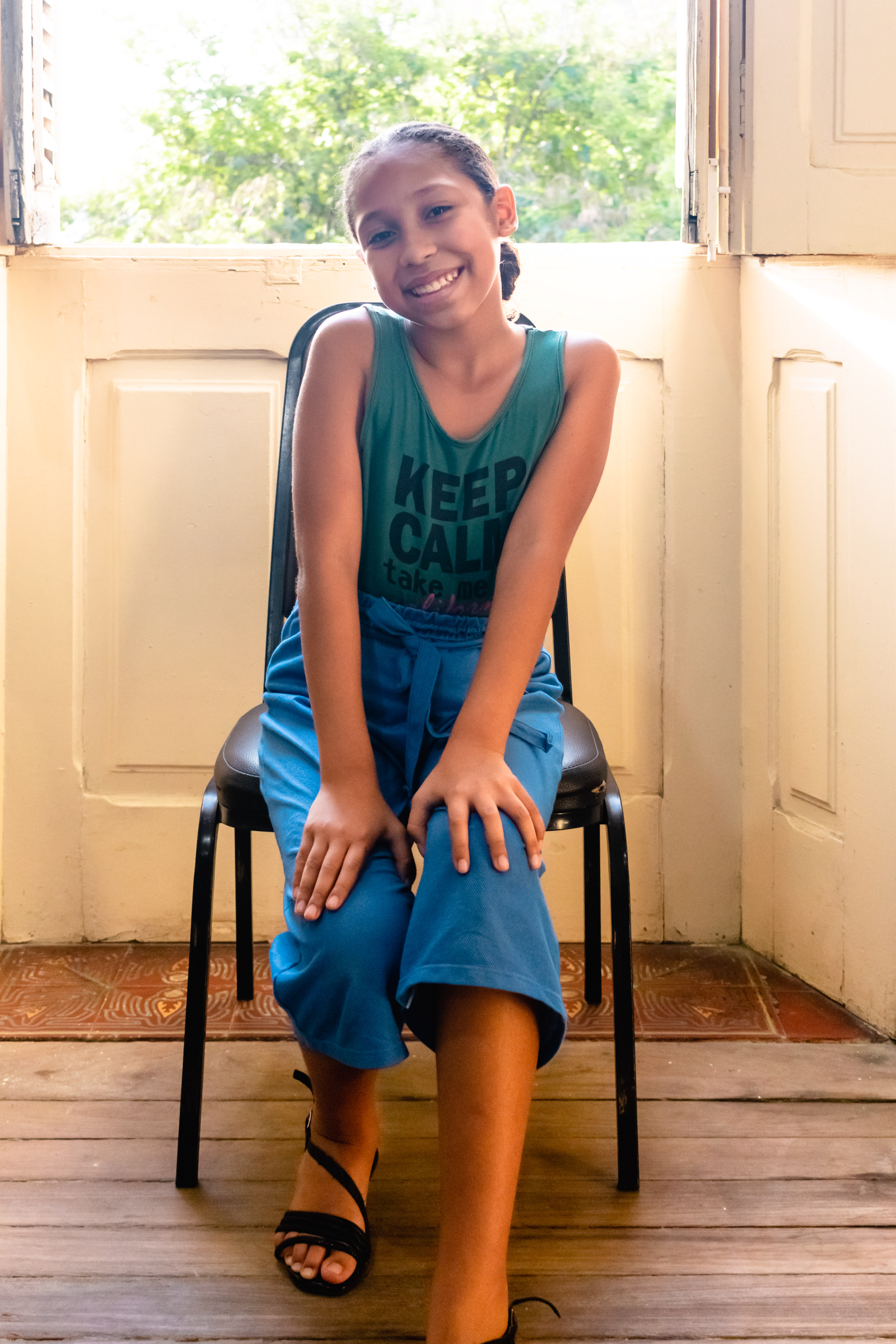 Josielen é uma criança negra, de 9 anos. Está sentada em uma cadeira em frente a uma janela e sorrindo para a câmera. 