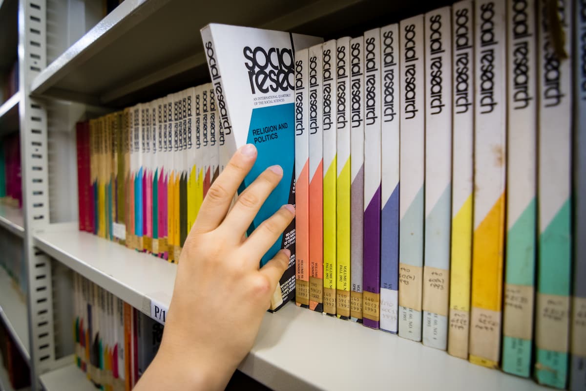 Fotografia mostra a mão de uma pessoa retirando um livro de uma estante com vários outros.