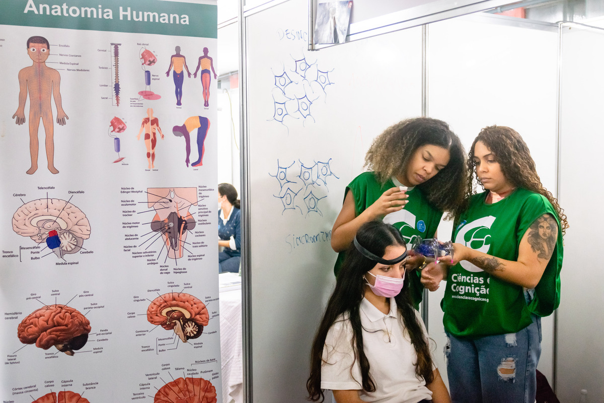 Fotografia do estande Ciências e Cognição. À direita, duas monitoras ajudam uma estudante a participar do experimento. À esquerda, há um cartaz com diversas representações da anatomia humana, com destaque para o cérebro.