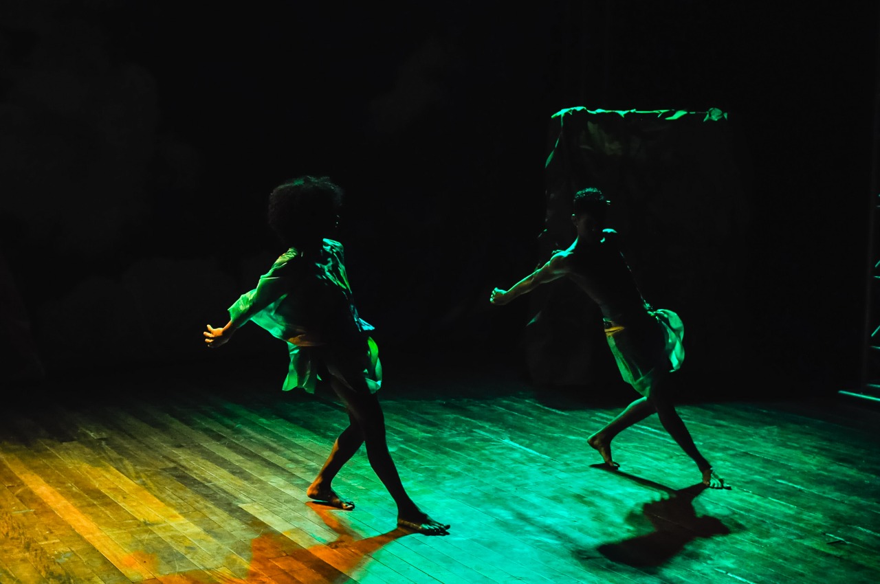 Foto na contraluz mostra dois corpos dançando na penumbra. A iluminação tem tons de verde.