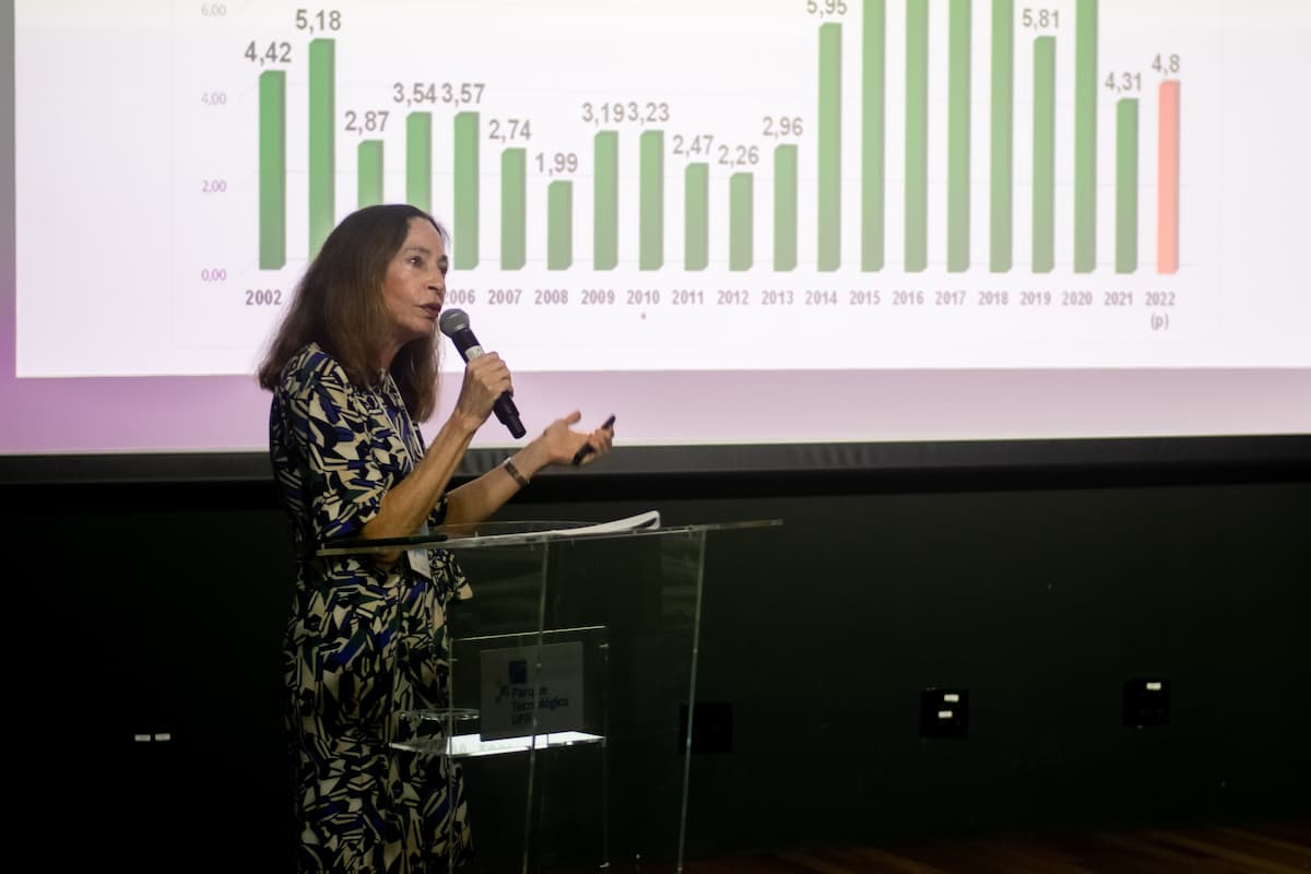 Fotografia mostra a professora Margarida Gutierrez, uma mulher branca de cabelos castanhos abaixo dos ombros, usando um vestido estampado branco, azul e preto, palestrando em um púlpito. Ao fundo, um slide mostra um gráfico em barra.