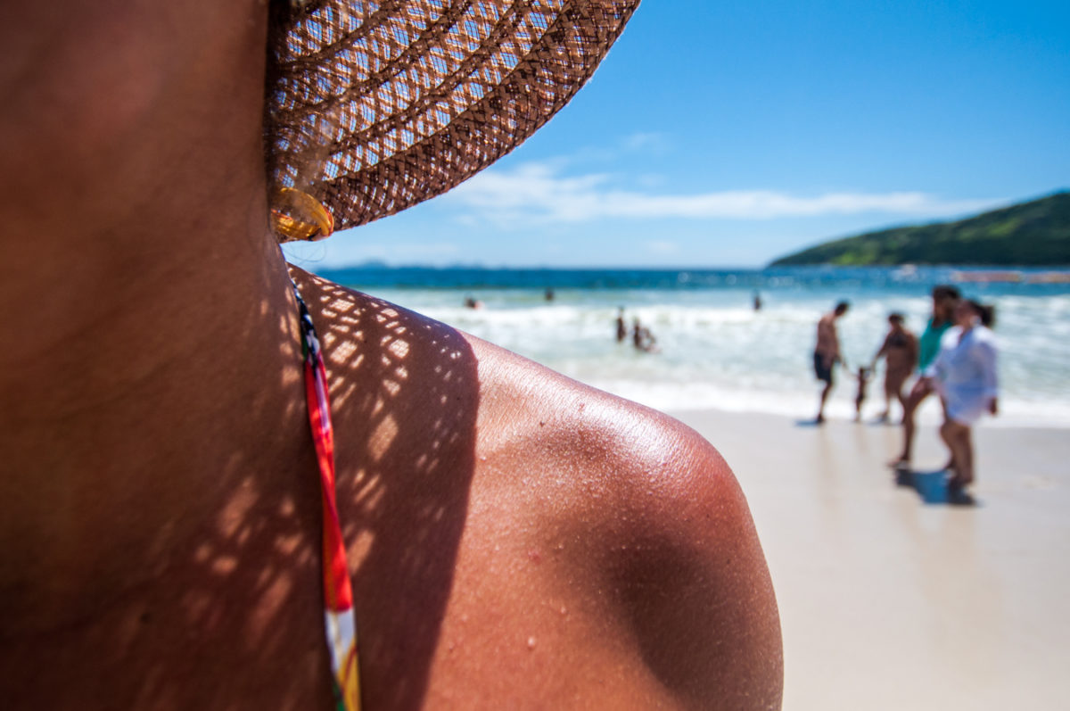 Fotografia mostra, em primeiro plano, uma mulher com ombro e pescoço à mostra, com chapéu e biquíni na praia. Ao fundo, pessoas caminham na areia e nadam no mar.