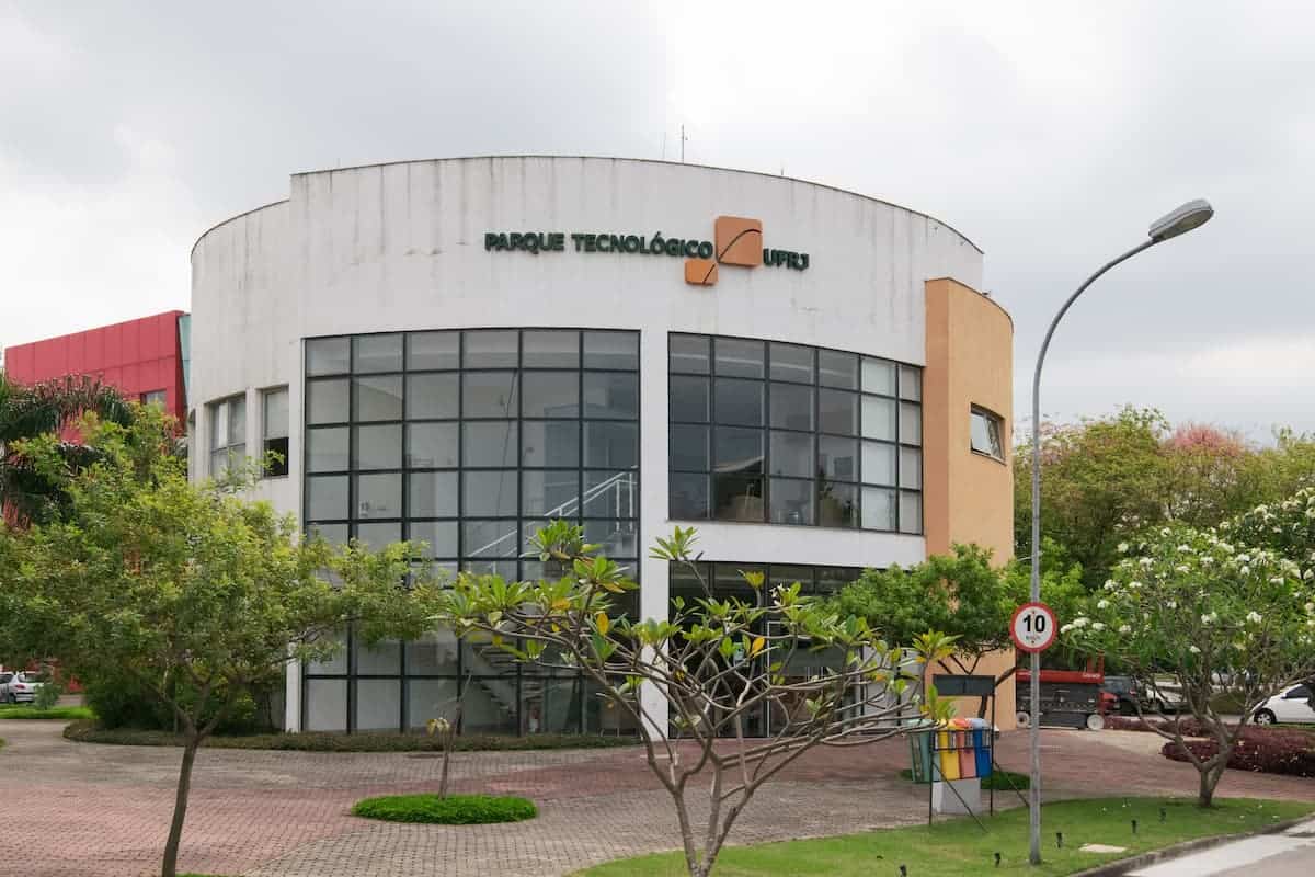 Fotografia mostra um prédio circular em branco, amarelo e laranja. No alto do prédio está o logo do Parque Tecnológico. A calçada do prédio tem várias árvores.
