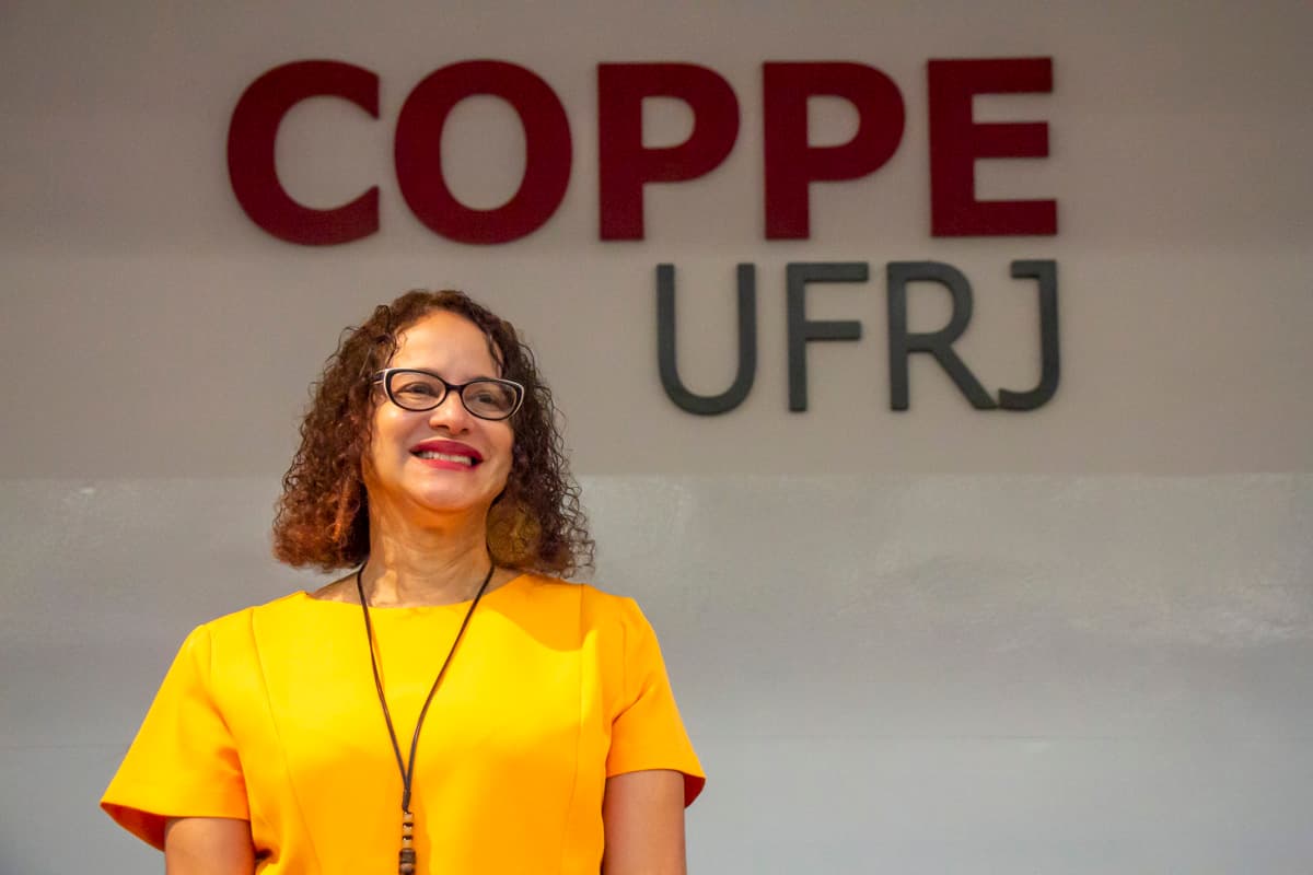 Fotografia mostra a ministra Luciana Santos, uma mulher negra, com cabelos cacheados na altura dos ombros. Luciana usa óculos, blusa amarela e sorri. Em segundo plano está o logo da Coppe UFRJ em vermelho.