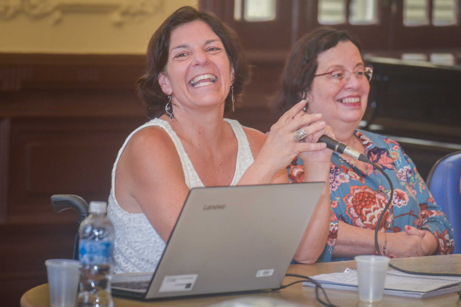 Fotografia de Anna Paula Feminella, mulher branca, de meia idade, que possui cabelos lisos na altura do pescoço e está vestindo uma camiseta branca. Ela está sorrindo e sentada a uma mesa em frente a um microfone e um notebook cinza. Ao seu lado encontra-se Amélia Rosalvo.