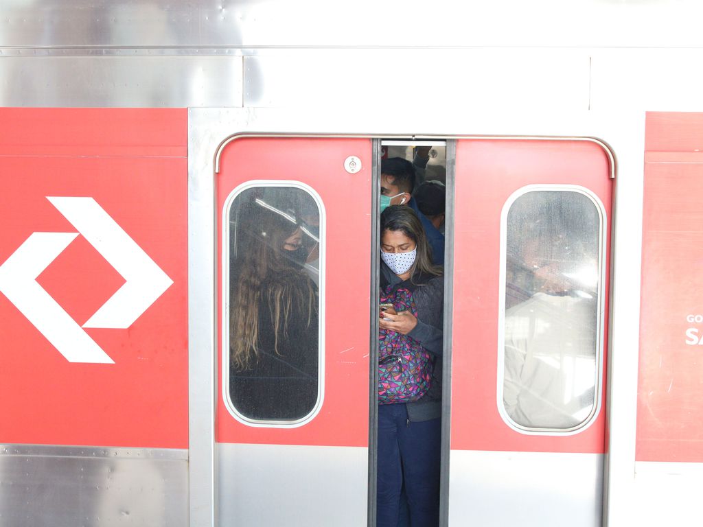 Fotografia mostra usuários utilizando máscaras em um trem paulista lotado. As portas vermelhas estão fechando enquanto uma mulher de máscara usa o celular.