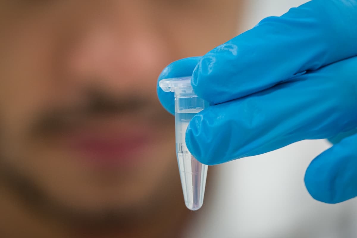 Fotografia mostra, em destaque, uma luva azul segurando um microtubo para centrífuga repleto de líquido transparente. Ao fundo, um homem observa a amostra.