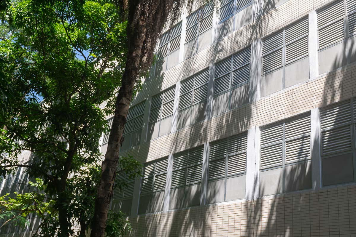 Foto em dia de sol da fachada da Residência Estudantil da UFRJ. Grandes árvores fazem sombra no prédio.