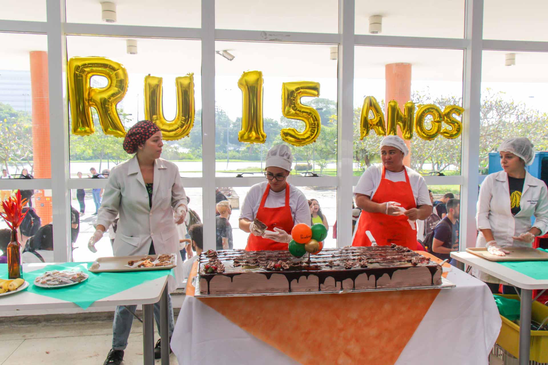 Foto de três funcionárias servindo um bolo de chocolate. Atrás delas, balões dourados apresentam a mensagem RU 15 ANOS