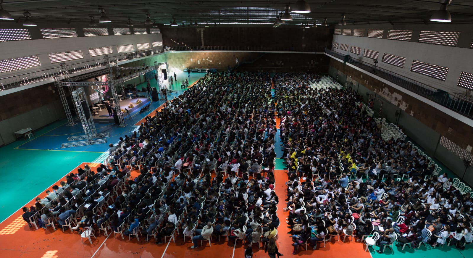 Fotografia de um grande auditório lotado, com pessoas sentadas em cadeiras enfileiradas em frente a um palco.