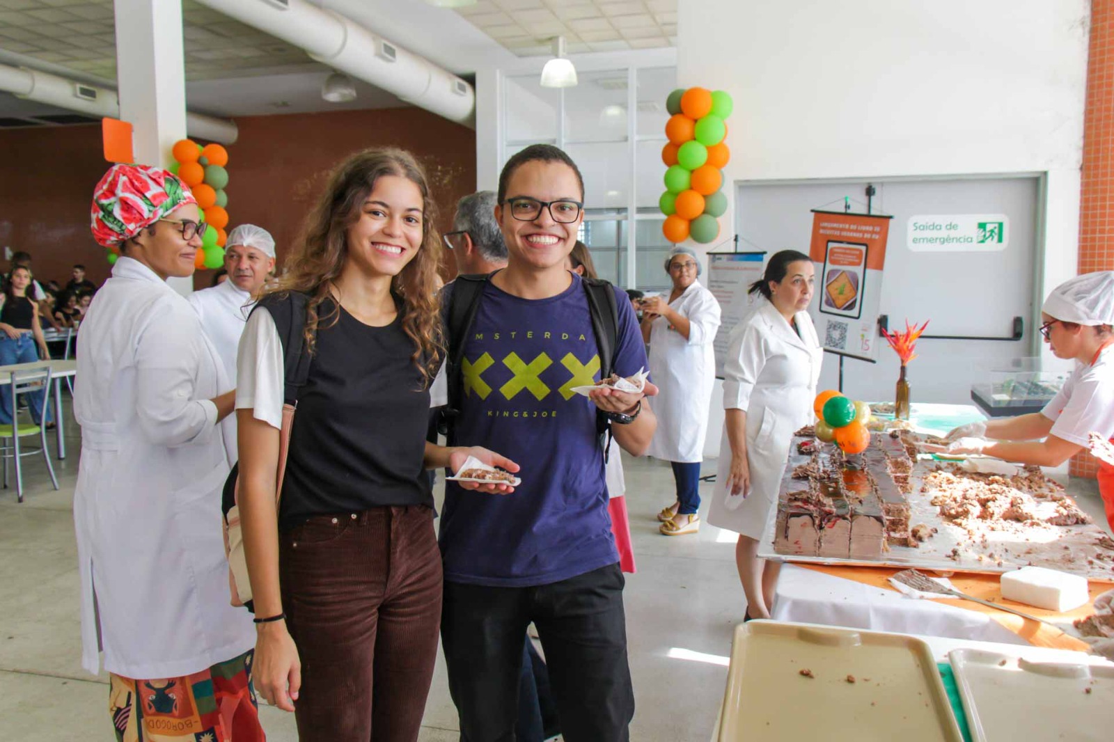 Dois alunos posam pra foto enquanto seguram bolo de chocolate. 