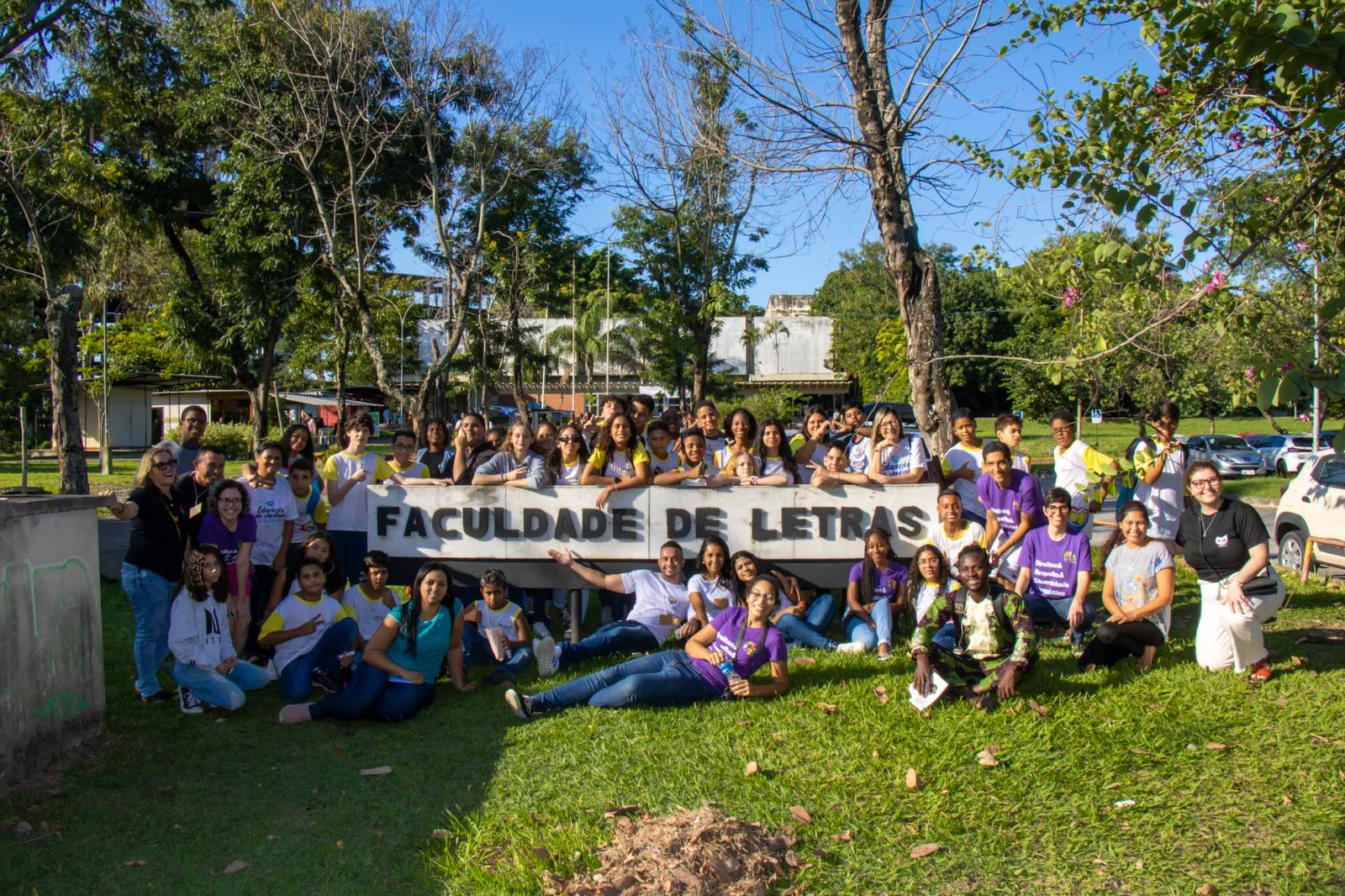 Fotografia ao ar livre em que dezenas de estudantes estão em volta de uma placa com o texto: Faculdade de Letras.