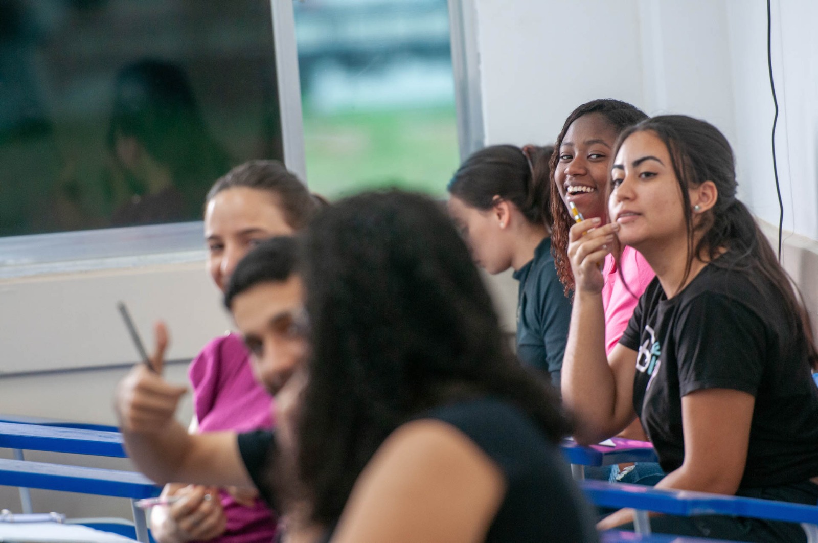 Descrição da imagem: Fotografia de seis estudantes sentados em carteiras escolares em uma sala de aula. Na primeira fileira, há duas mulheres e um homem, que faz sinal de positivo para a câmera. Atrás estão três estudantes, mulheres, sorrindo. Duas são brancas e uma negra.