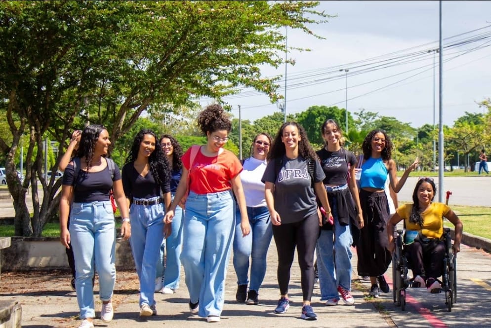 Descrição da imagem: Um grupo de dez estudantes caminha por um campus da UFRJ. Todas são mulheres e estão sorrindo. A maioria é negra e uma delas usa cadeira de rodas.