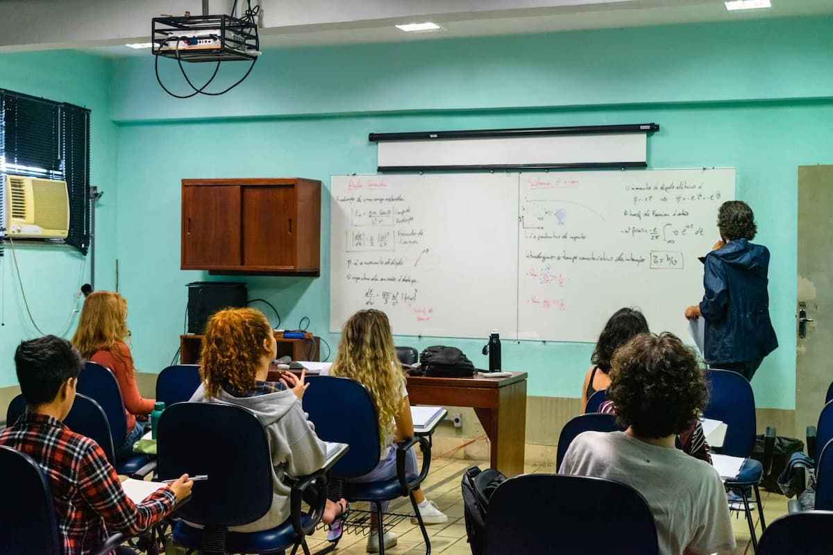Fotografia mostra uma sala de aula. Ao fundo, um professor, de costas, escreve fórmulas matemáticas em um quadro branco. Espalhados pela sala, alunos assistem à aula.