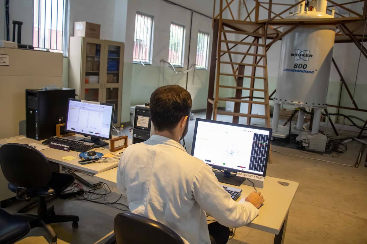 Fotografia mostra um pesquisador de costas mexendo no computador. Ao fundo, uma escada leva para o topo de um espectômetro, uma grande máquina cilíndrica. 