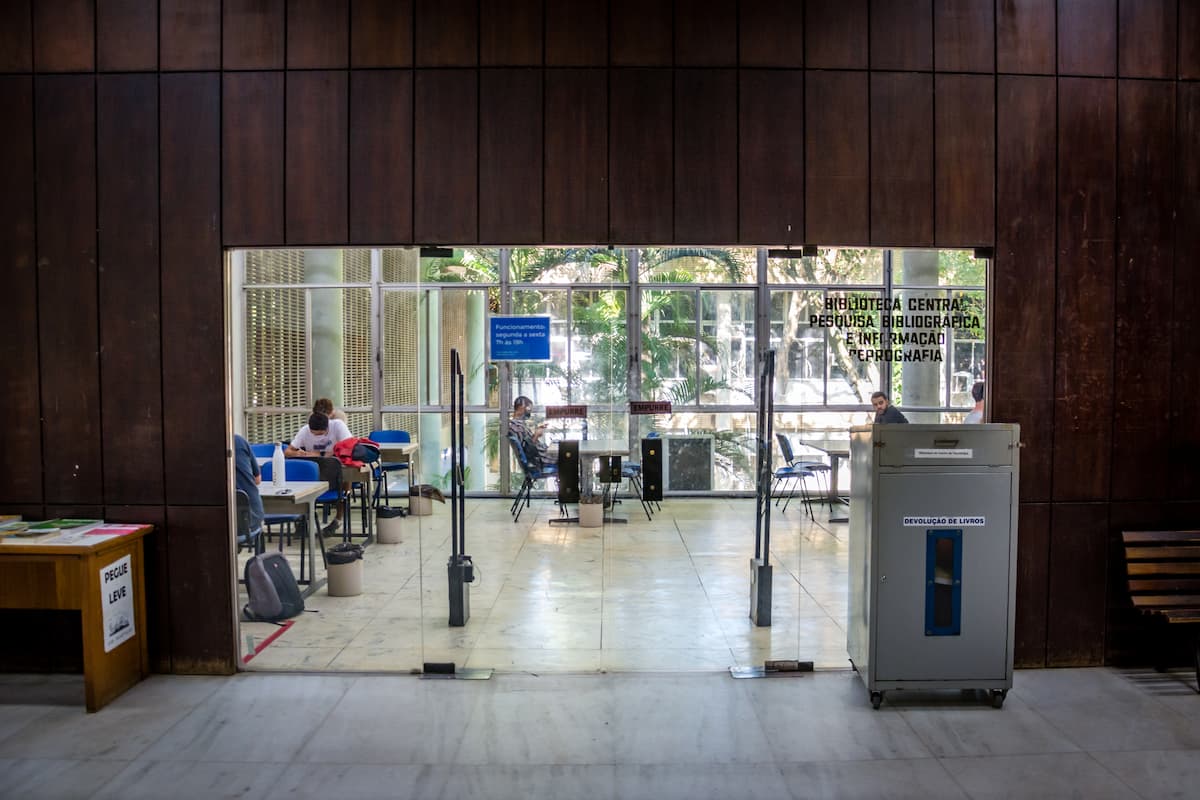 Fotografia do hall de entrada da biblioteca do CT. Em primeiro plano está uma parede de madeira que emoldura a porta de vidro que dá acesso ao espaço. Através do vidro, é possível ver estudantes em mesas.