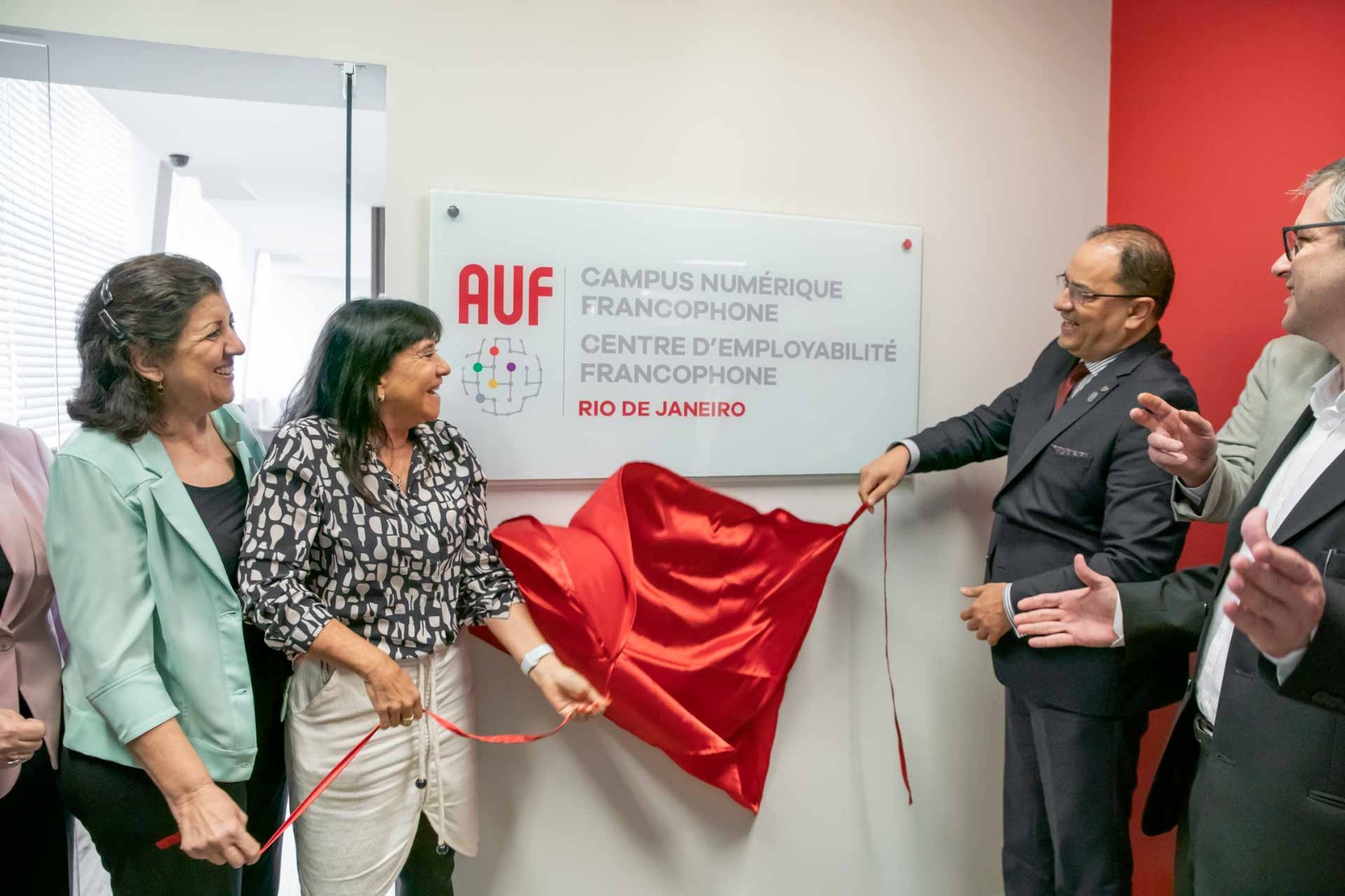 Duas mulheres.e dois homens retiram tecido vermelho da placa de inauguração da AUF, onde se lê, em francês: Campus Numerique Francophone | Centre Démployabilité Francophone Rio de Janeiro