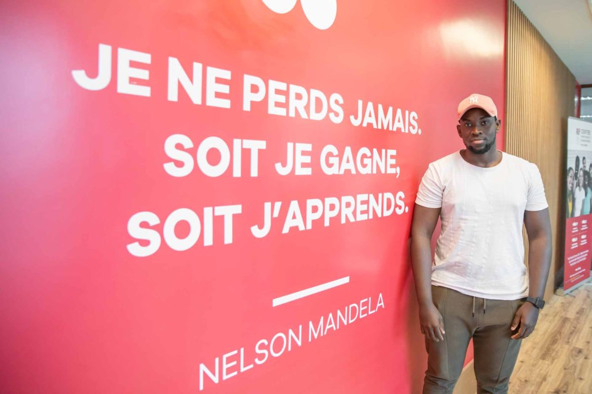 Aluno negro de boné posa ao lado de uma parede vermelha que contém frase de Nelson Mandela em francês, que significa: Eu não perco nunca. Ou eu ganho, ou eu aprendo. 