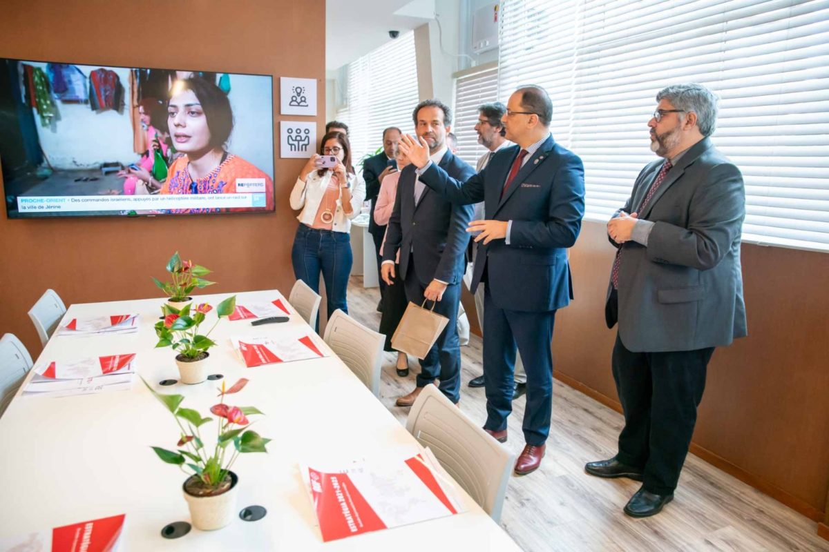 Pessoas estão em sala multimídia onde há uma televisão e uma mesa para reuniões, com plantas sobre ela. 