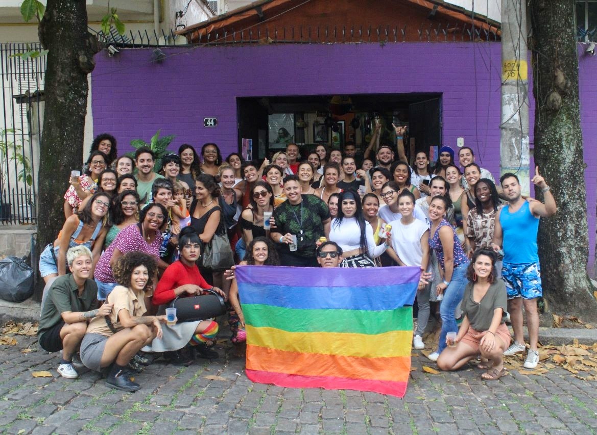 Várias pessoas posam para uma foto em frente a uma casa roxa. Na frente, elas seguram a bandeira dos movimentos LGBT+ om várias cores