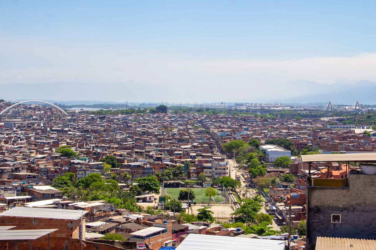 Em dia de céu claro, fotografia tirada do alto mostra a visão geral de diversas casas, um campo de futebol e uma rua larga em uma das favelas da Maré. Ao fundo se veem a Baía de Guanabara às margens da favela e a Cidade Universitária.