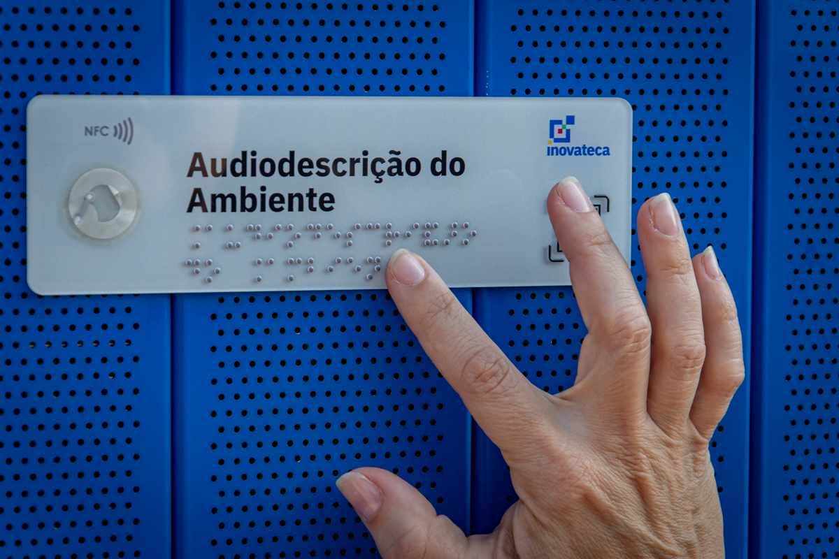 Fotografia de uma pequena placa em uma das paredes da Inovateca. O objeto indica que a audiodescrição do ambiente pode ser feita por Braille e NFC. Um usuário aproxima a mão para realizar a leitura tátil.