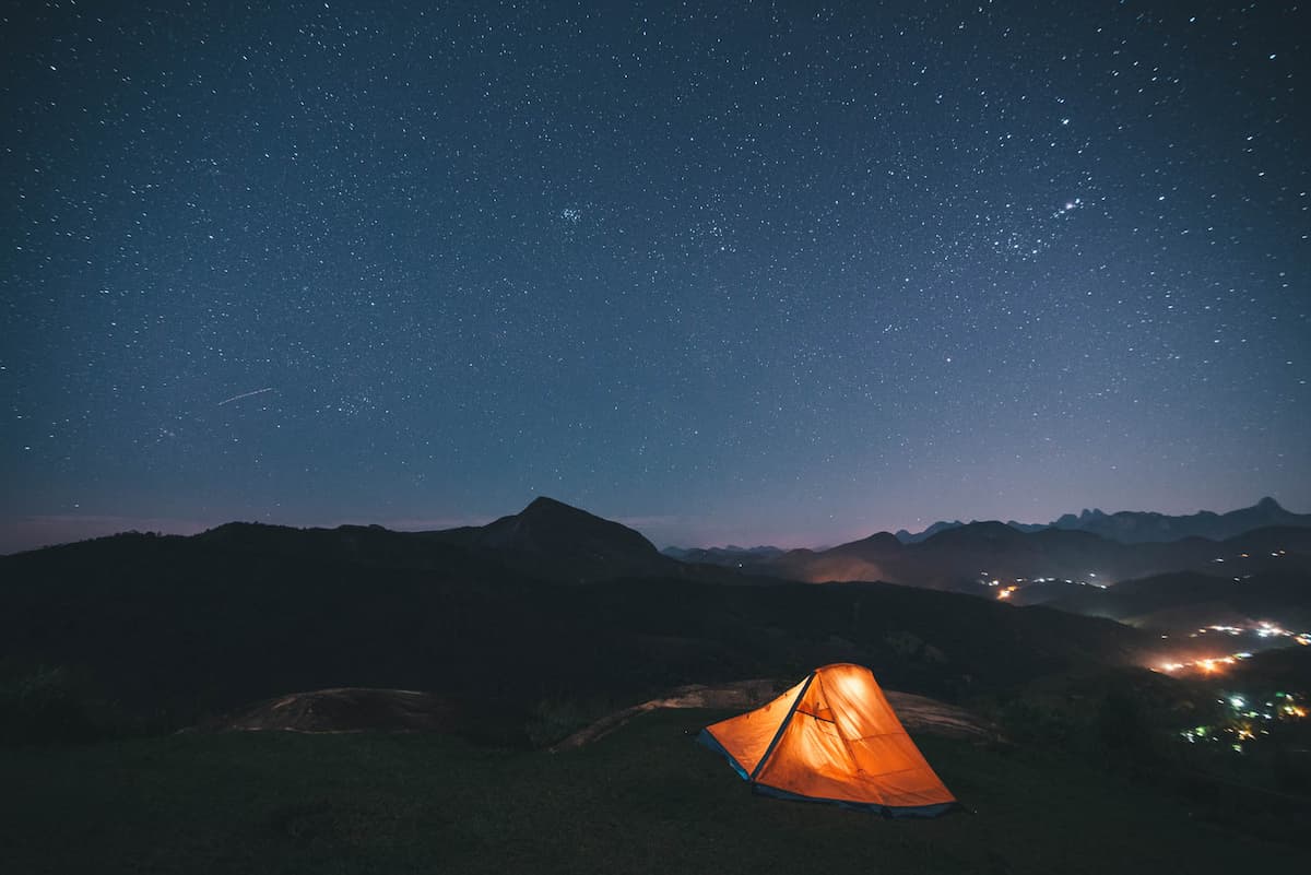 Fotografia mostra céu estrelado em meio a uma cadeia de montanhas. Em destaque, há uma barraca de acampamento amarela iluminada por uma luz na parte interna.