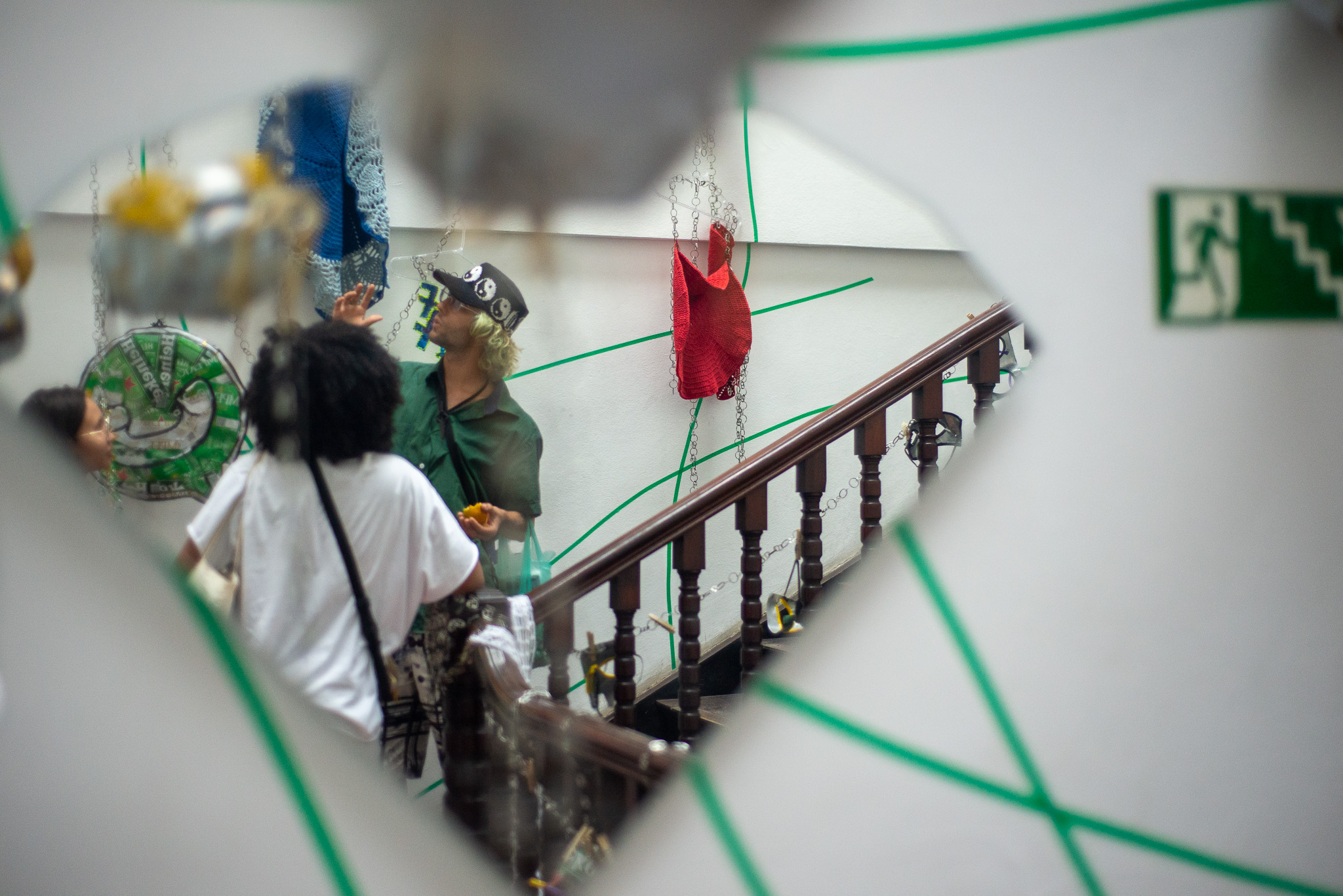 Um espelho quebrado reflete parte da instalação "MetaHall", no qual aparece uma escada e duas pessoas refletidas.