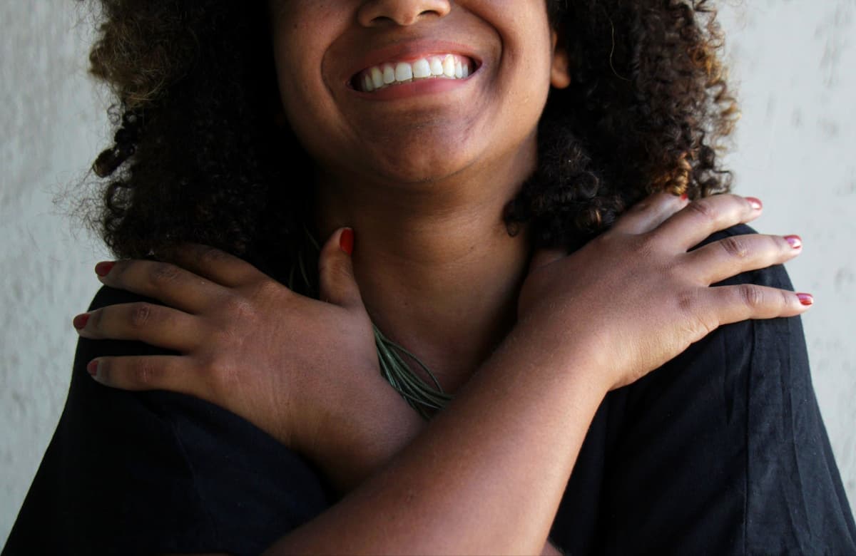 Fotografia de uma mulher negra sorrindo com destaque para região do busto. As mãos da mulher estão cruzadas sobre o peito em um gesto de abraço.