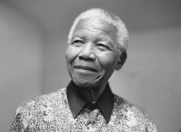 Fotografia em preto e branco de Nelson Mandela. O enquadramento é dos ombros para cima. Ele veste uma camisa estampada e sorri suavemente.