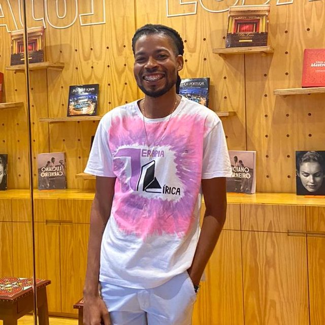 Um jovem negro de barba veste calça branca e camisa branca com rosa. Ele posa para a foto sorrindo. Ao fundo, paredes e móveis de madeira. 