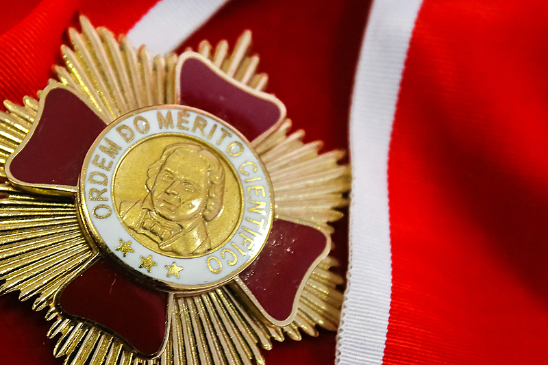 Fotografia da medalha do mérito científico, com detalhes em dourado e vermelho. Sobre um um tecido também vermelho com uma listra branca.
