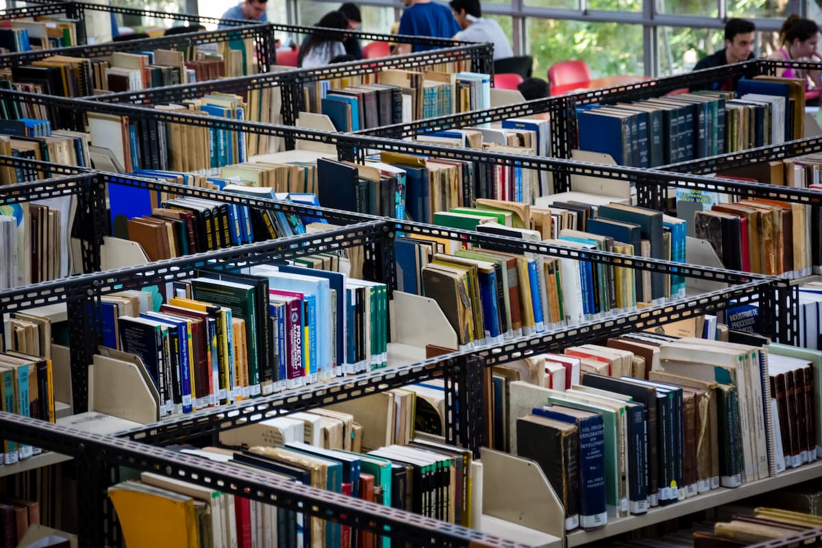 Fotografia da Biblioteca do Centro de Tecnologia, onde há diversos corredores de estantes cheias de livros. Na parte superior da imagem, pessoas estudam nas mesas da biblioteca.
