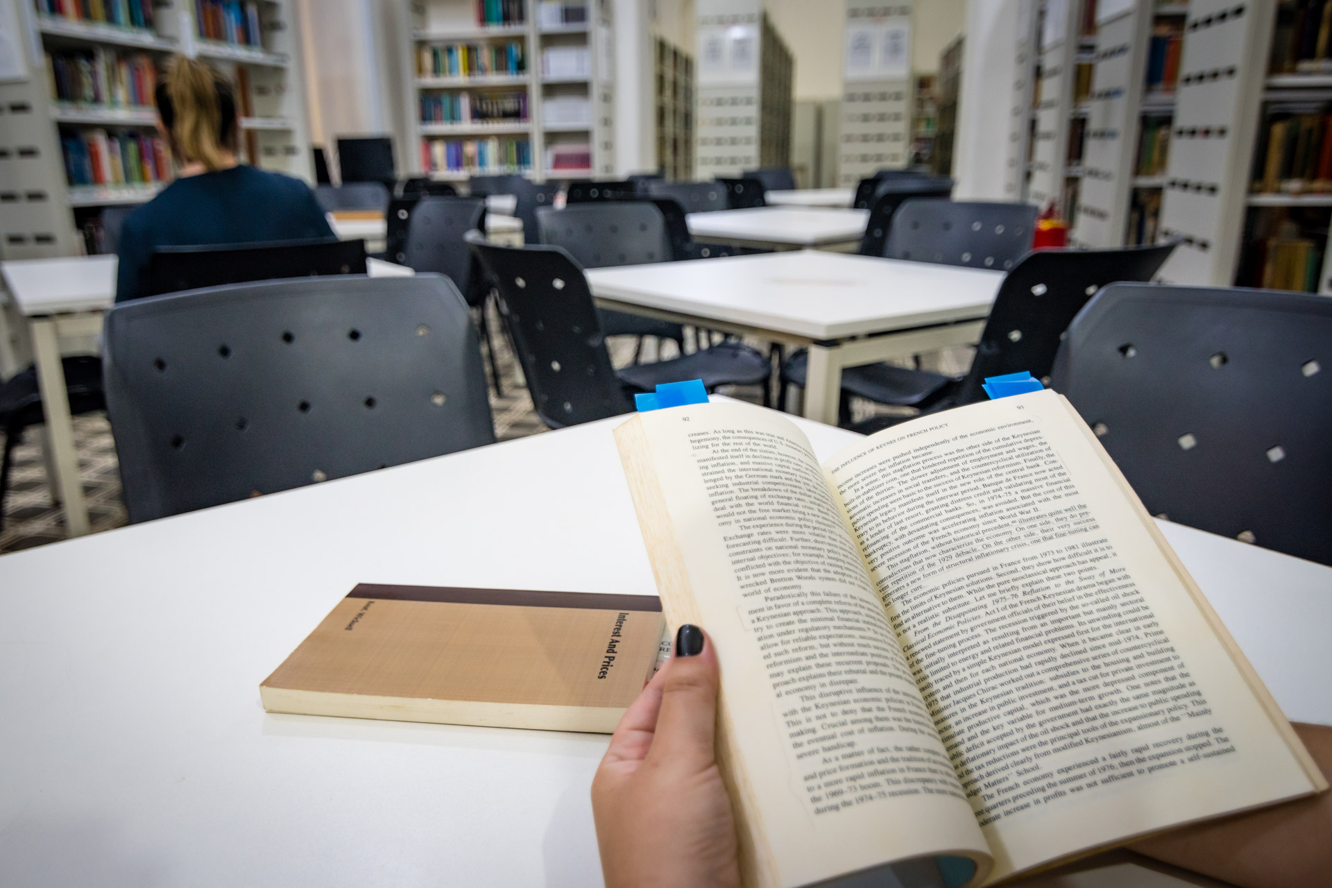 Fotografia de uma biblioteca onde, em destaque, uma pessoa segura um livro aberto enquanto outro exemplar está em cima de uma mesa. Ao fundo, mesas, cadeiras e prateleiras com livros.