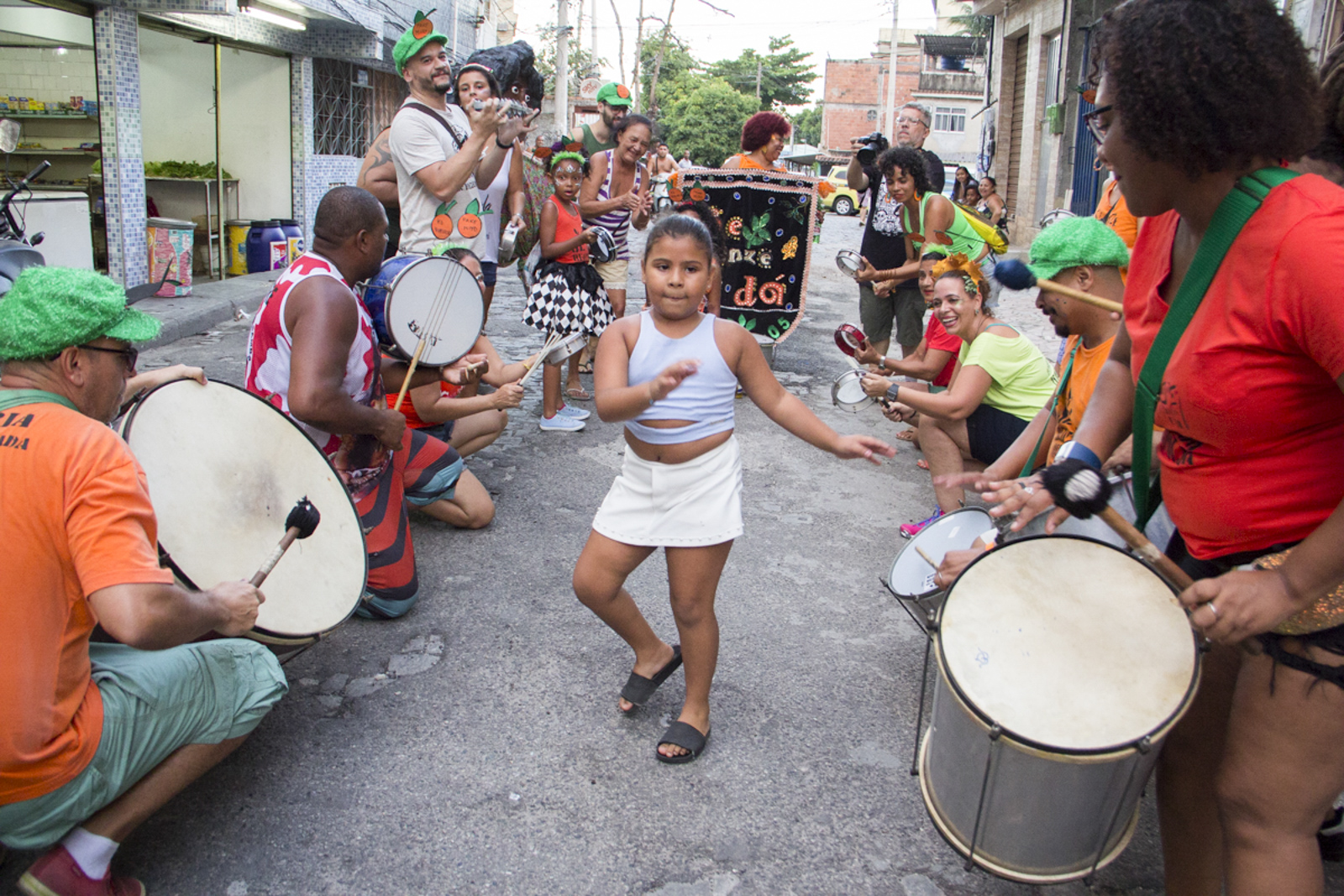 Uma criança samba em uma rua enquanto pessoas com tambores e outros instrumentos tocam ao redor dela.