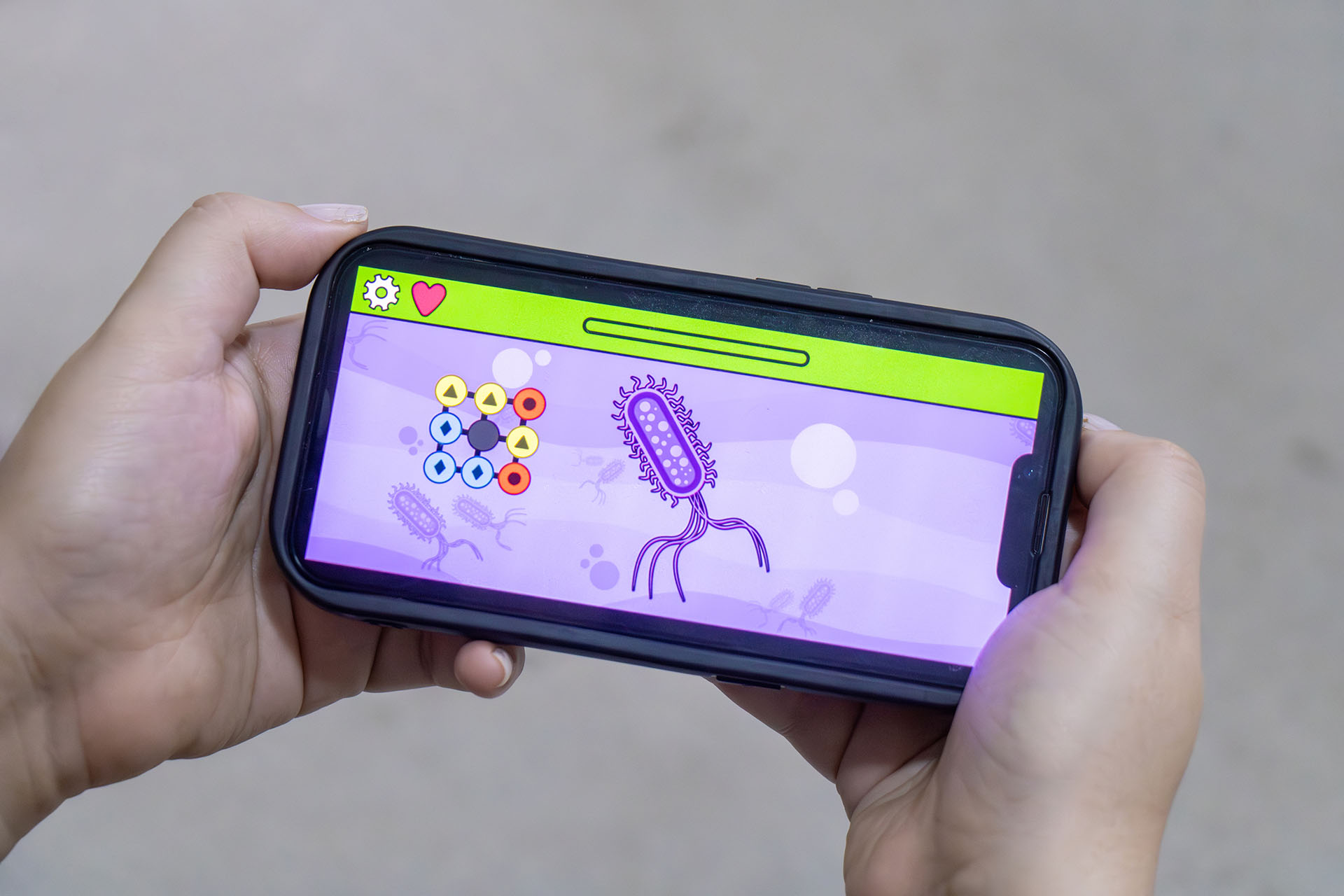 Fotografia mostra as mãos de uma pessoa segurando um celular na horizontal. O aparelho mostra captura de tela do jogo DiscoveRX em que o usuário precisa construir uma molécula capaz de combater uma bactéria.