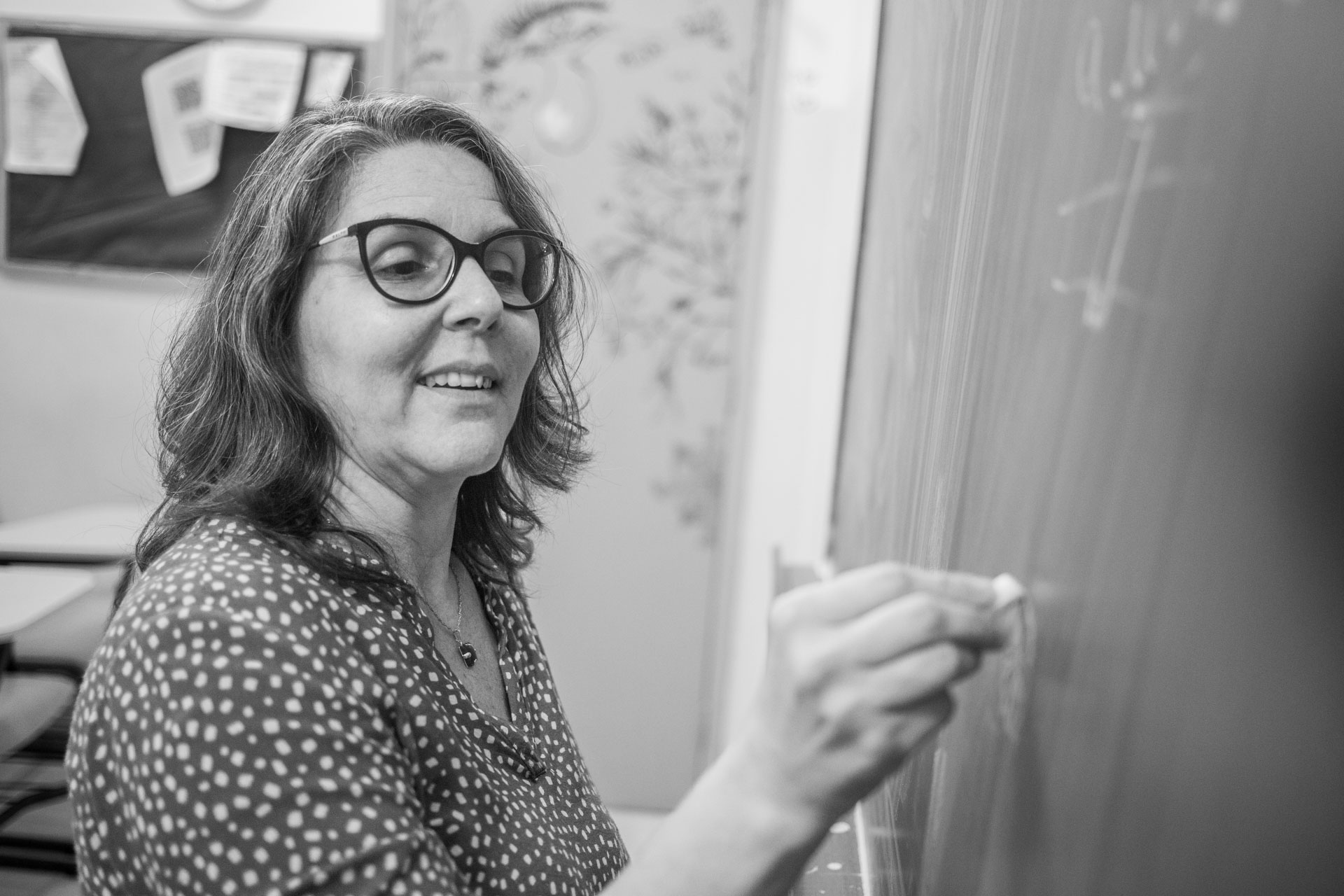 Foto em preto e branco de professora escrevendo com giz em quadro negro. Ela está sorrindo e tem cabelos lisos soltos até a altura dos ombros.