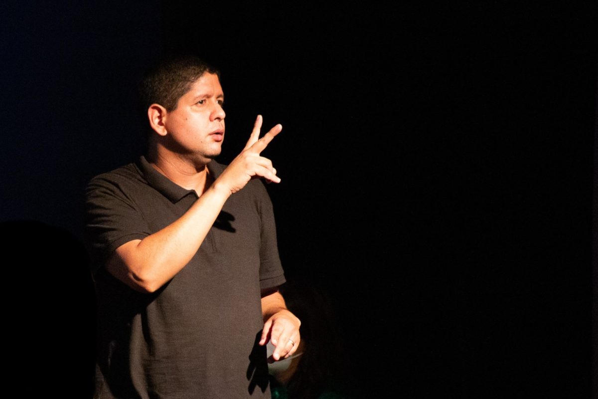 Fotografia de um intérprete de Libras, no momento de uma sinalização, de pele branca e cabelos curtos e escuros. Ele está vestindo uma camisa polo preta.
