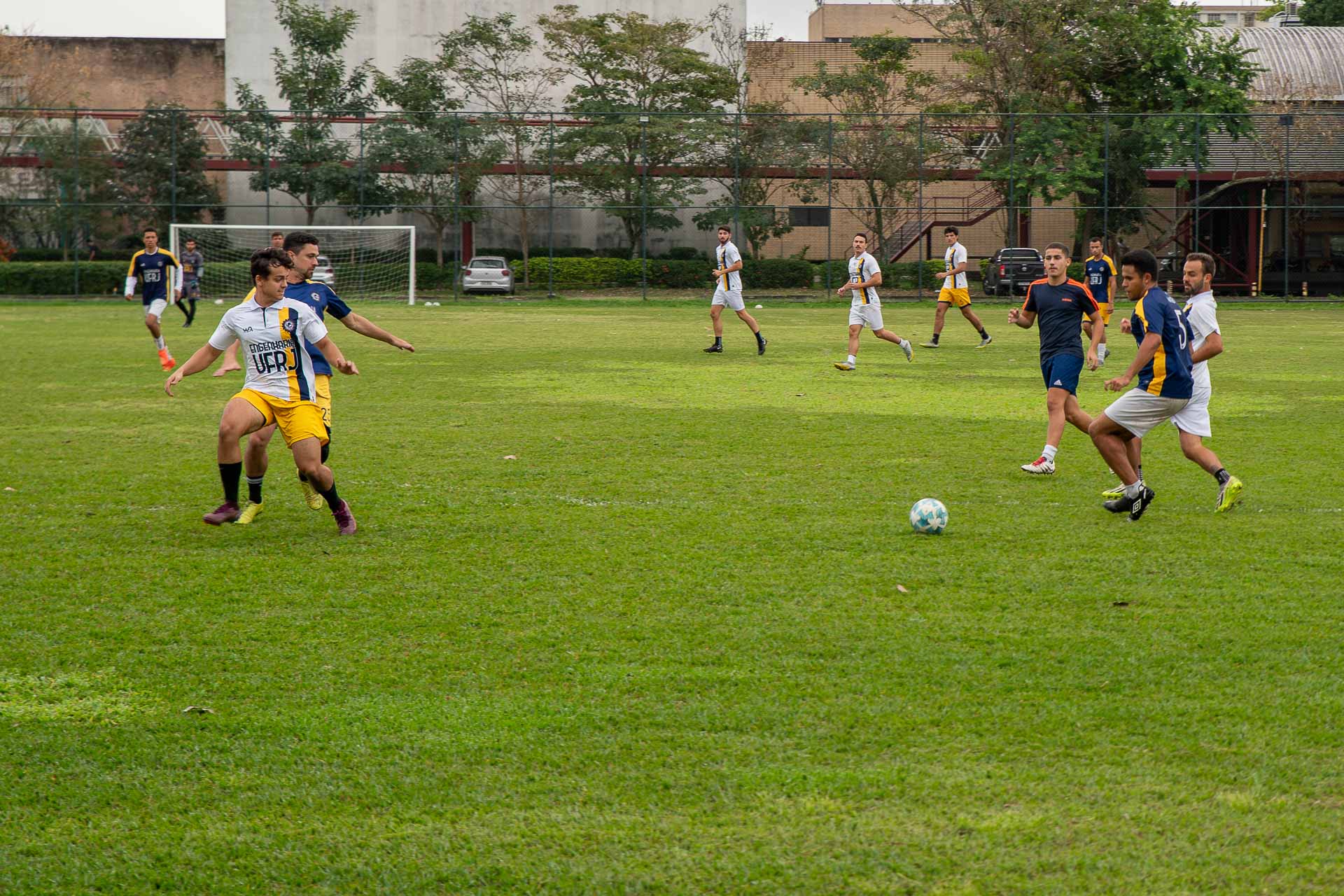 Fotografia mostra grupo de estudantes jogando futebol em um campo. Um dos times usa uniforme branco e amarelo, o outro usa uniforme azul escuro e amarelo, ambos com os dizeres Engenharia UFRJ.