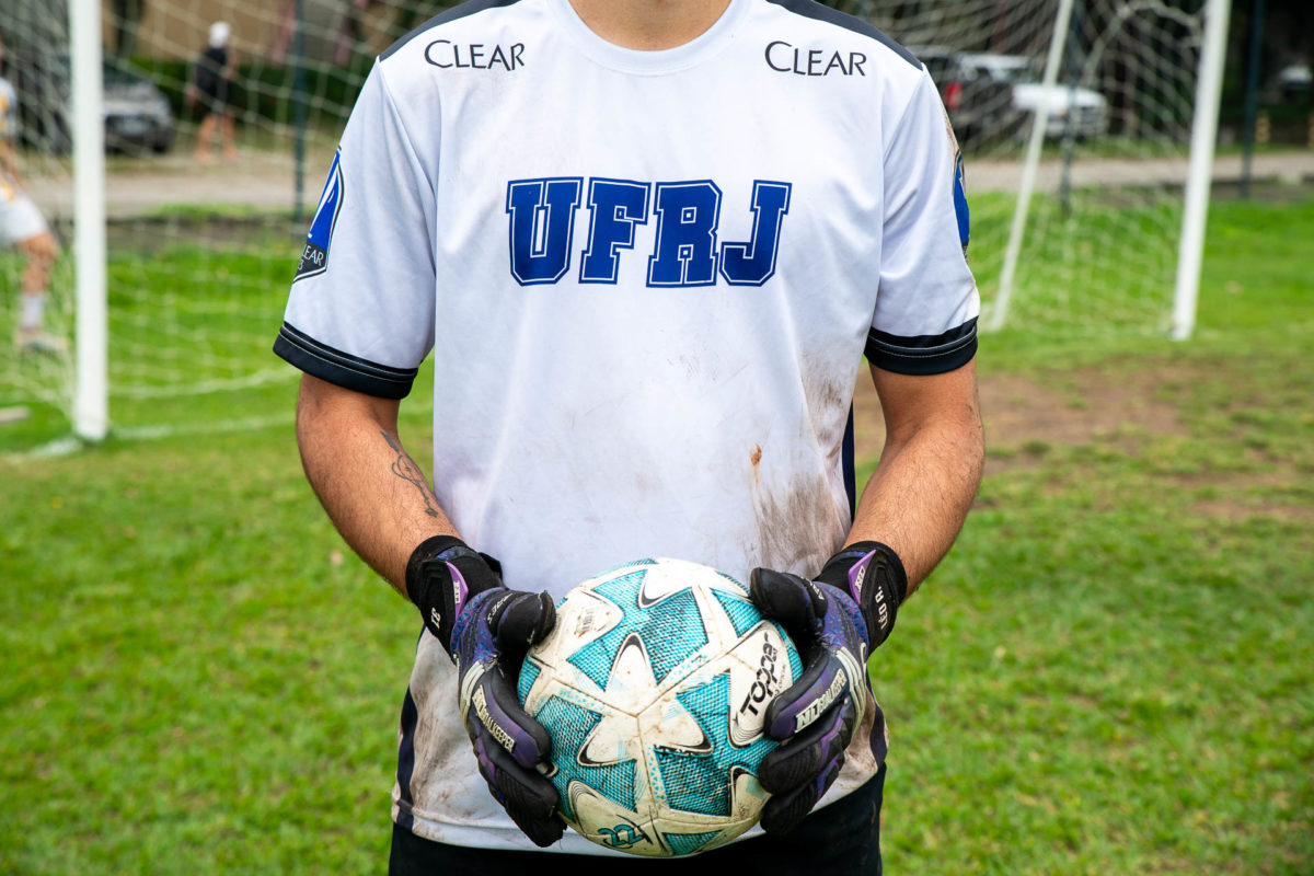 Fotografia mostra o torso de um estudante branco que veste um uniforme esportivo sujo de terra com a logomarca da UFRJ. Ele está com luvas de goleiro e segura uma bola. No fundo da imagem há uma baliza de futebol 