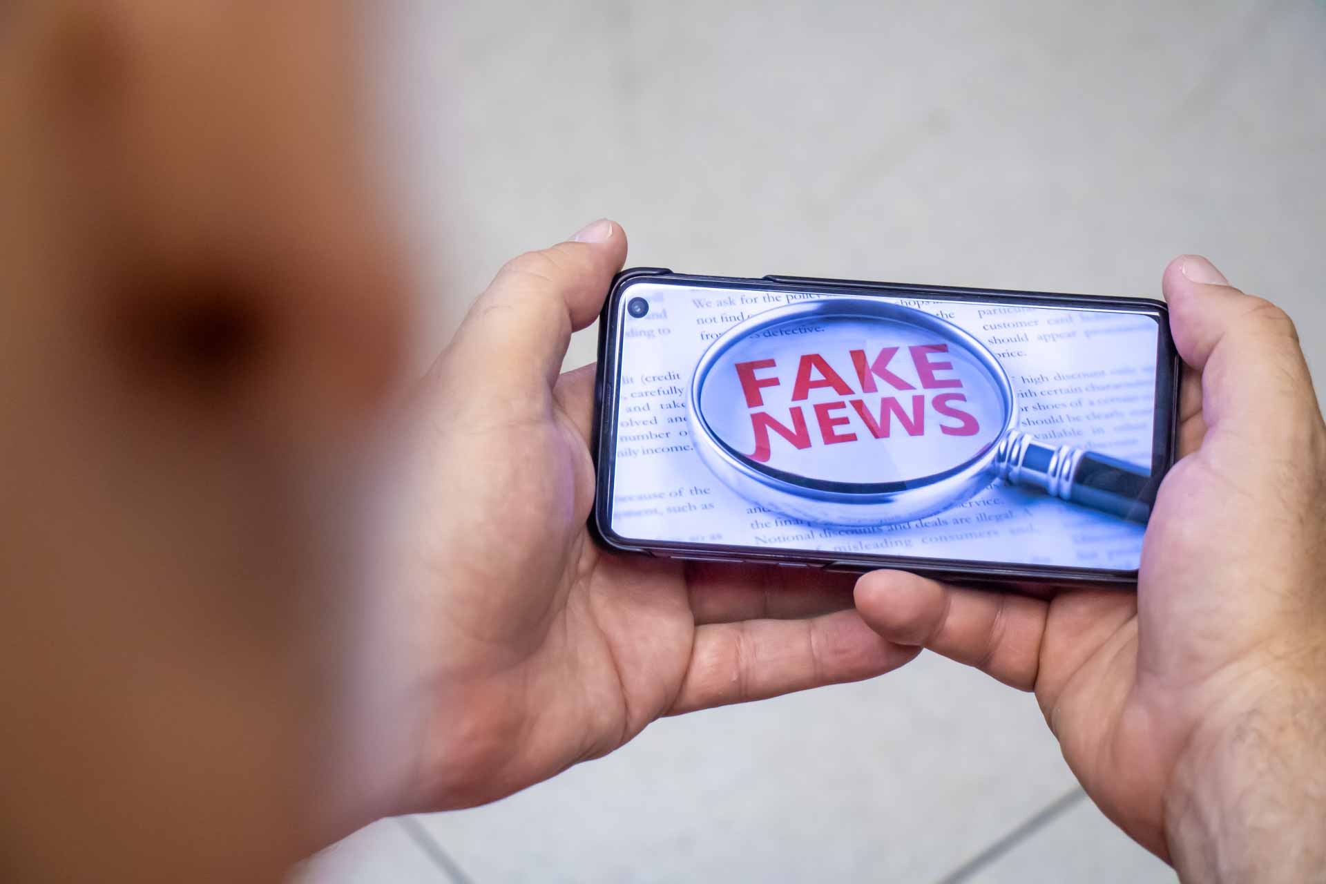 Mãos segurando celular no qual pode-se ver a imagem de uma lupa sobre as palavras "fake news".