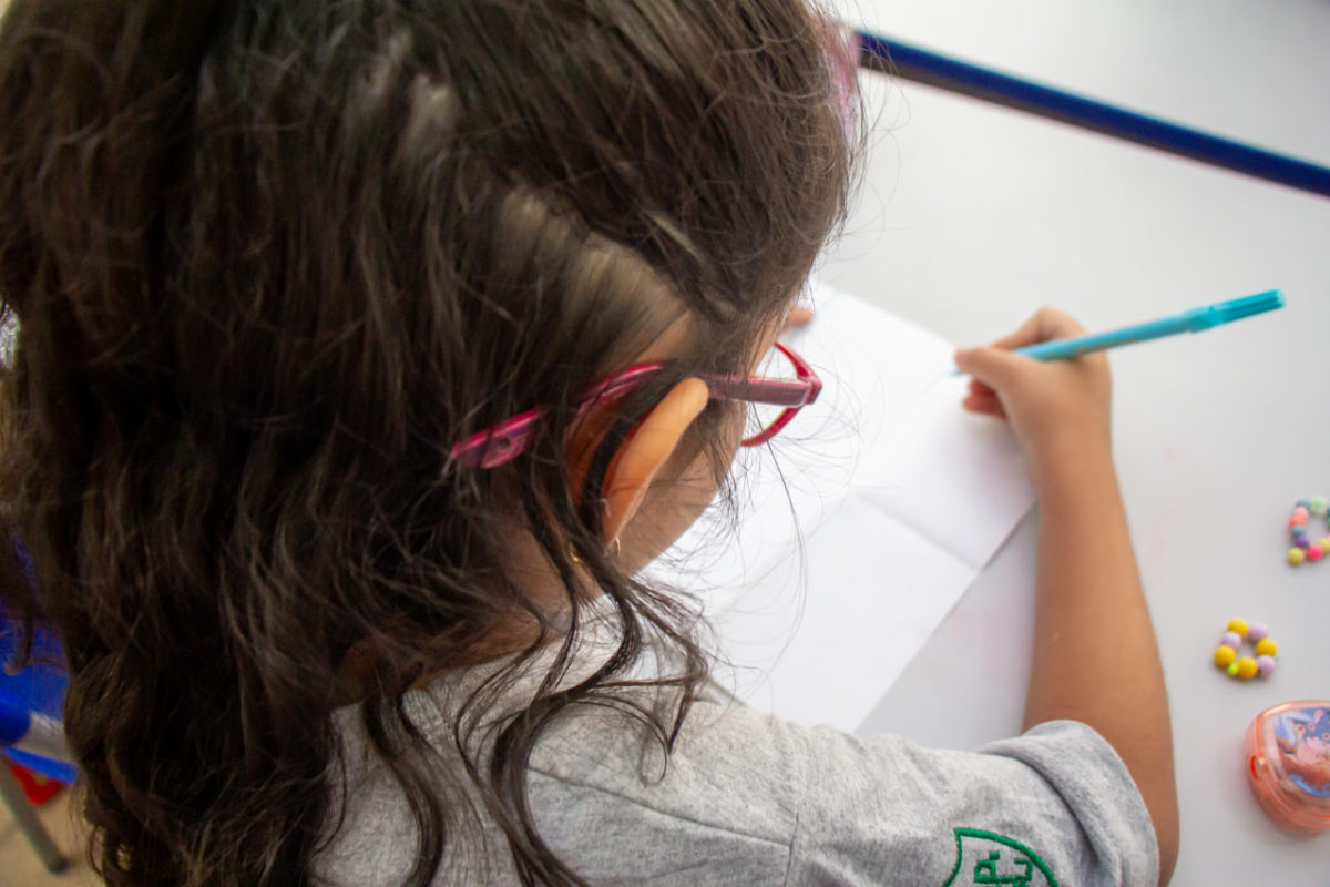 De cima, fotografia registra criança segurando uma caneta hidrocor azul enquanto desenha em folha de papel apoiada na mesa. Ela usa óculos vermelhos e tem cabelos longos castanhos.
