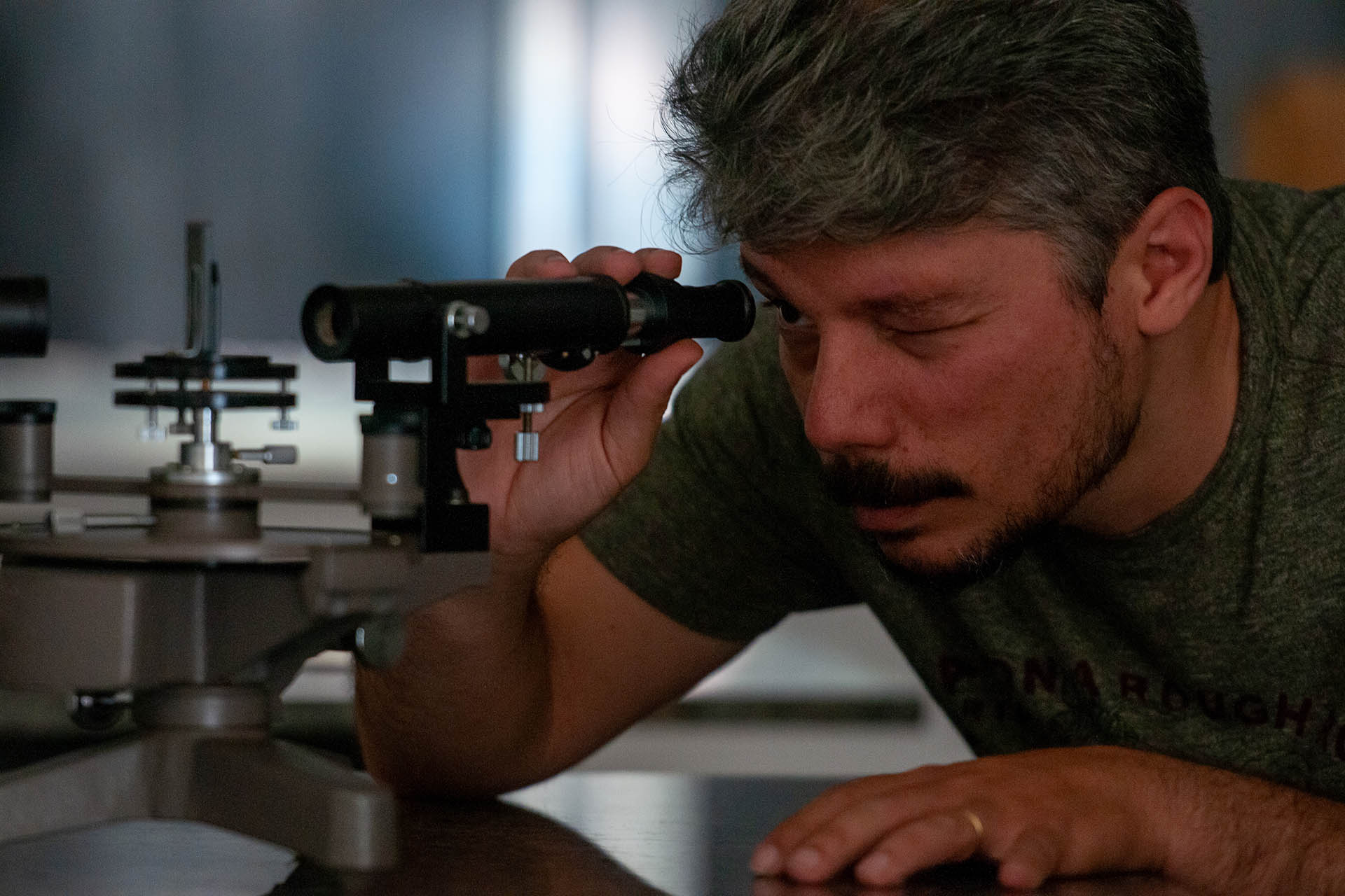 Fotografia de um homem branco, que possui cabelos grisalhos, bigode e barba rala, olhando através de um microscópio. Ele o segura com a mão direita enquanto apoia a mão esquerda sobre a mesa.