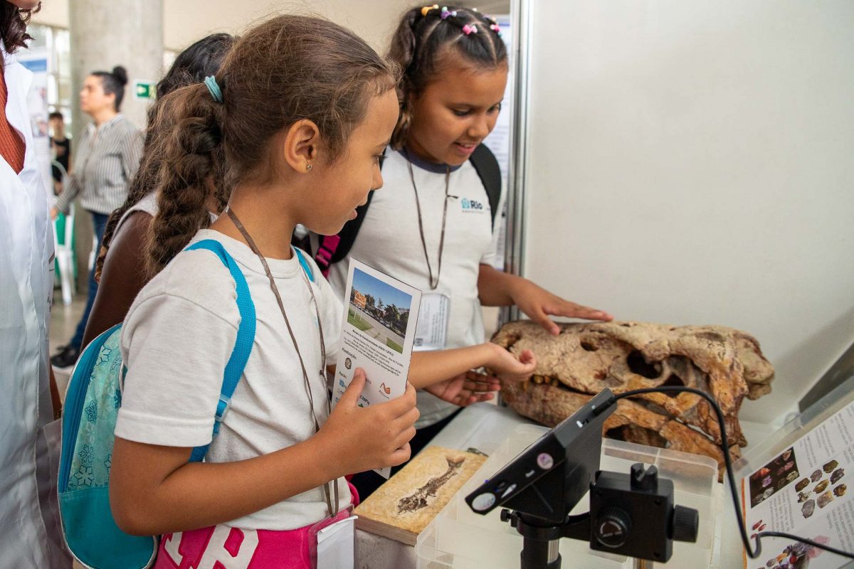 Fotografia de duas crianças interagindo com objetos de paleontologia na 20ª Semana Nacional de Ciência e Tecnologia (SNCT). Elas usam uniforme escolar e mochila e tocam um fóssil da cabeça de um dinossauro.
