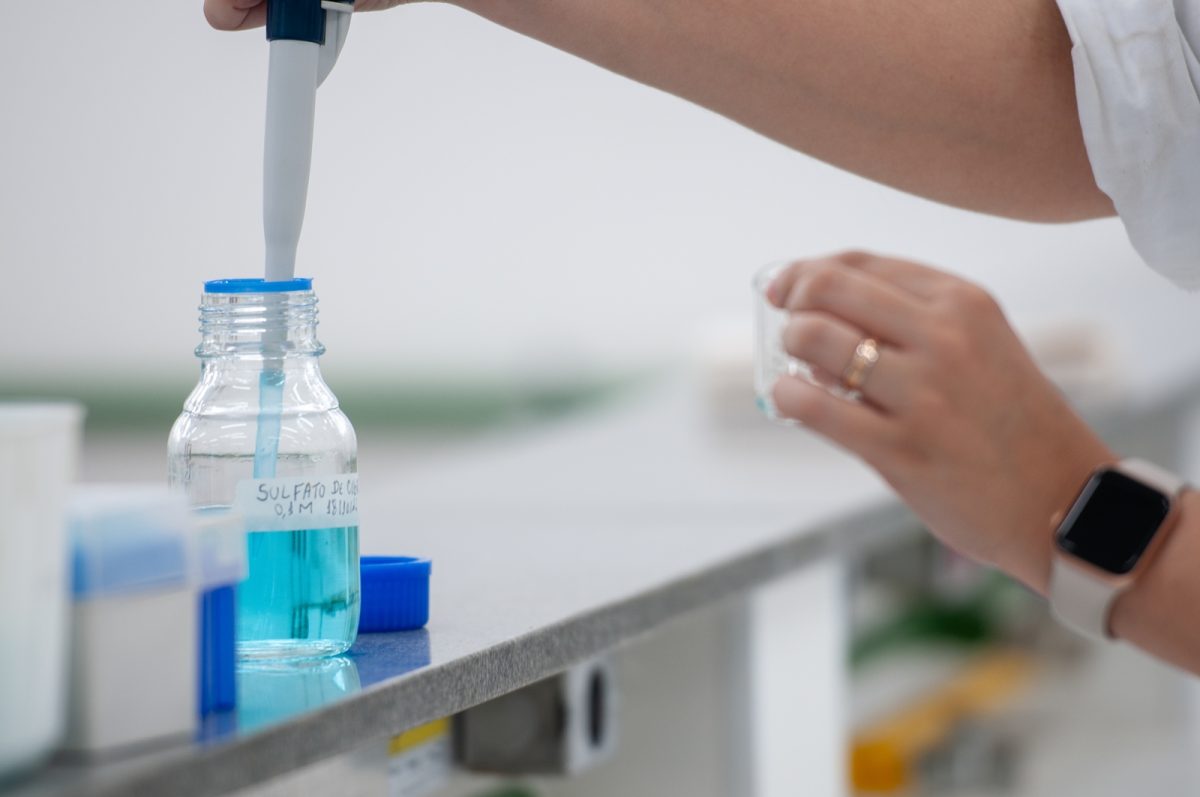 Fotografia em plano fechado dentro de um laboratório, captura o momento em que uma pessoa manipula uma espécie de seringa em um frasco transparente contendo um líquido azul.