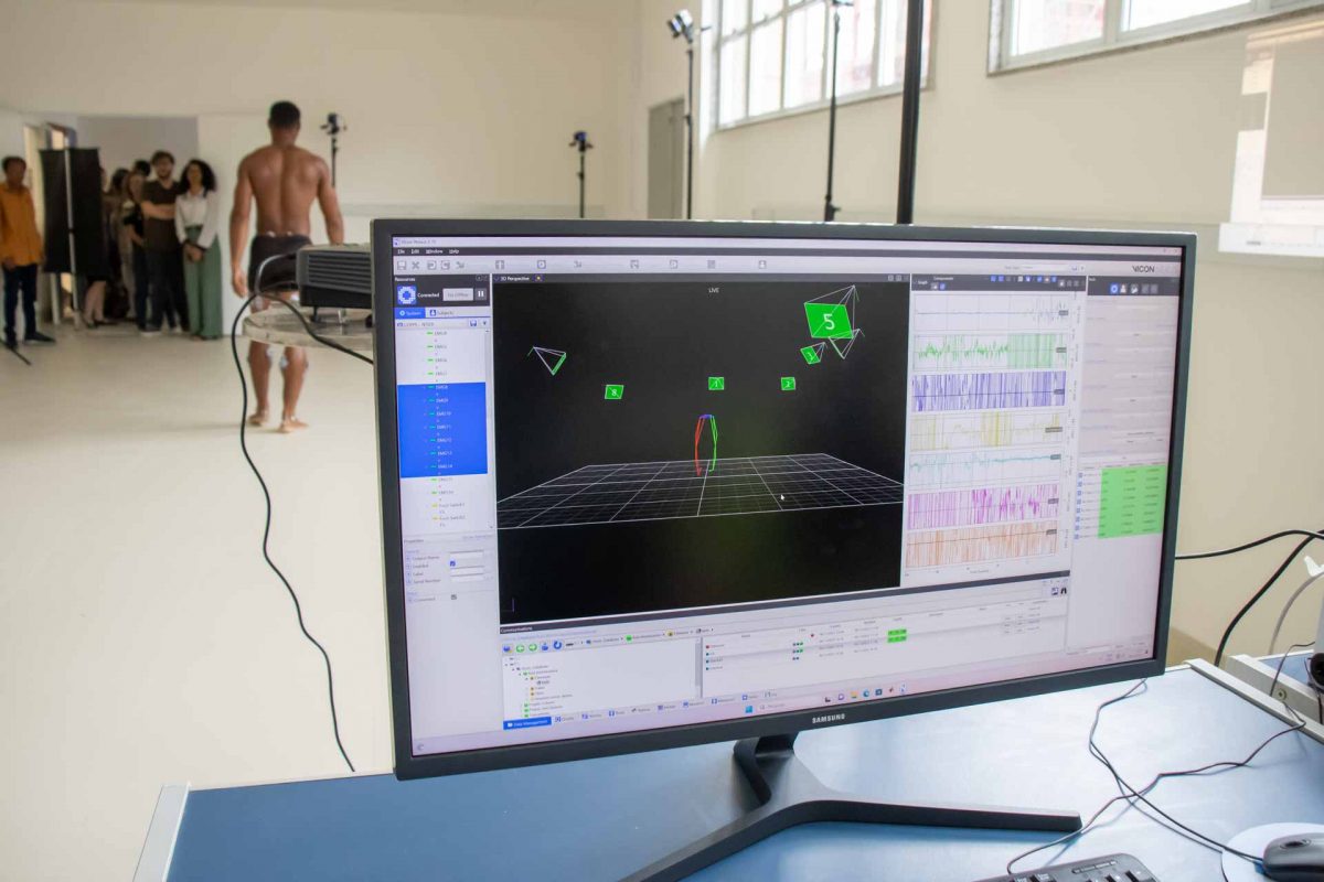 Fotografia mostra, em primeiro plano, um computador com gráfico. Ao fundo, um homem de short está com sensores de movimento pelo corpo. 